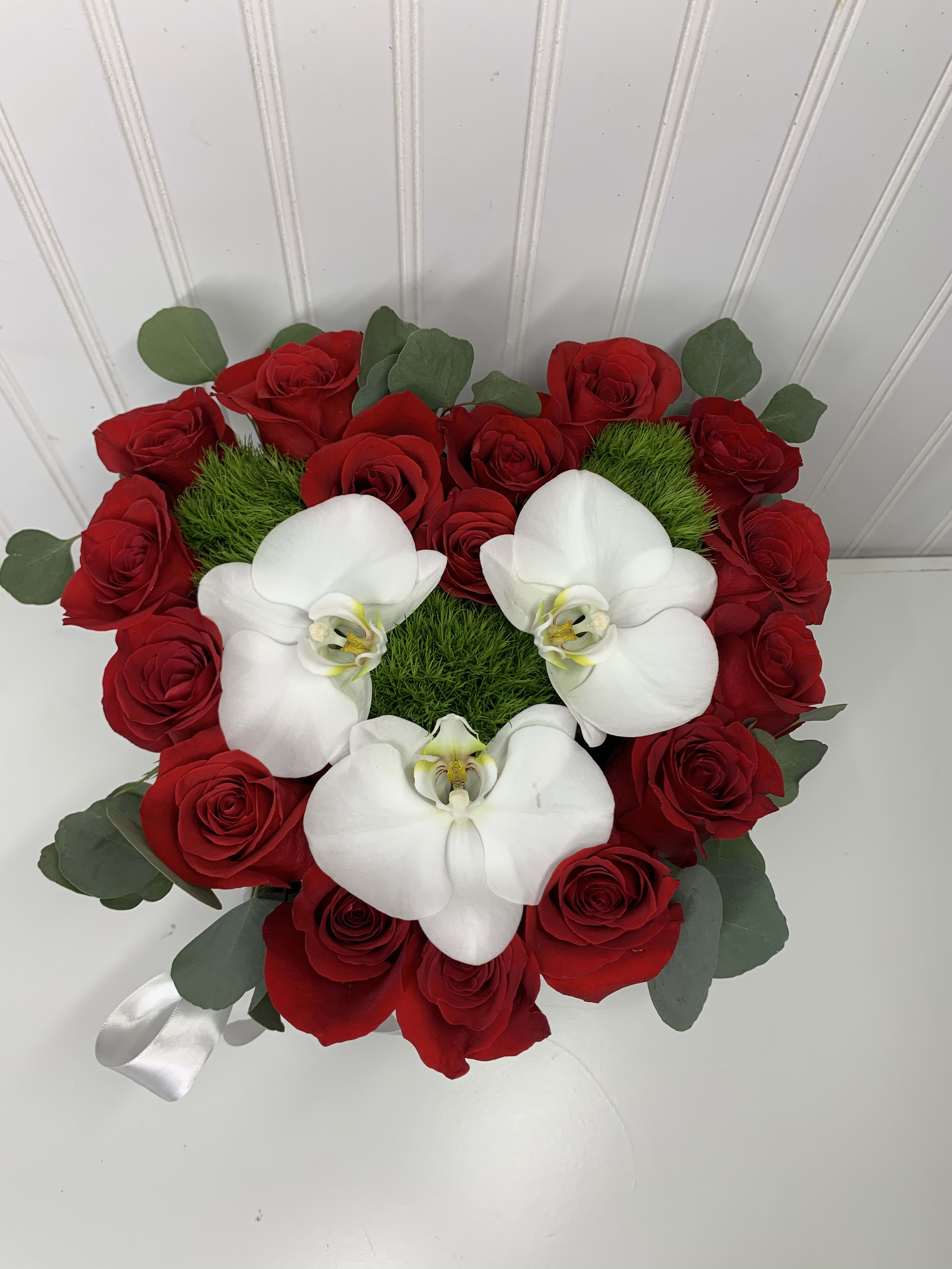 Heart-shaped arrangement of red roses with white orchids and green moss accents.