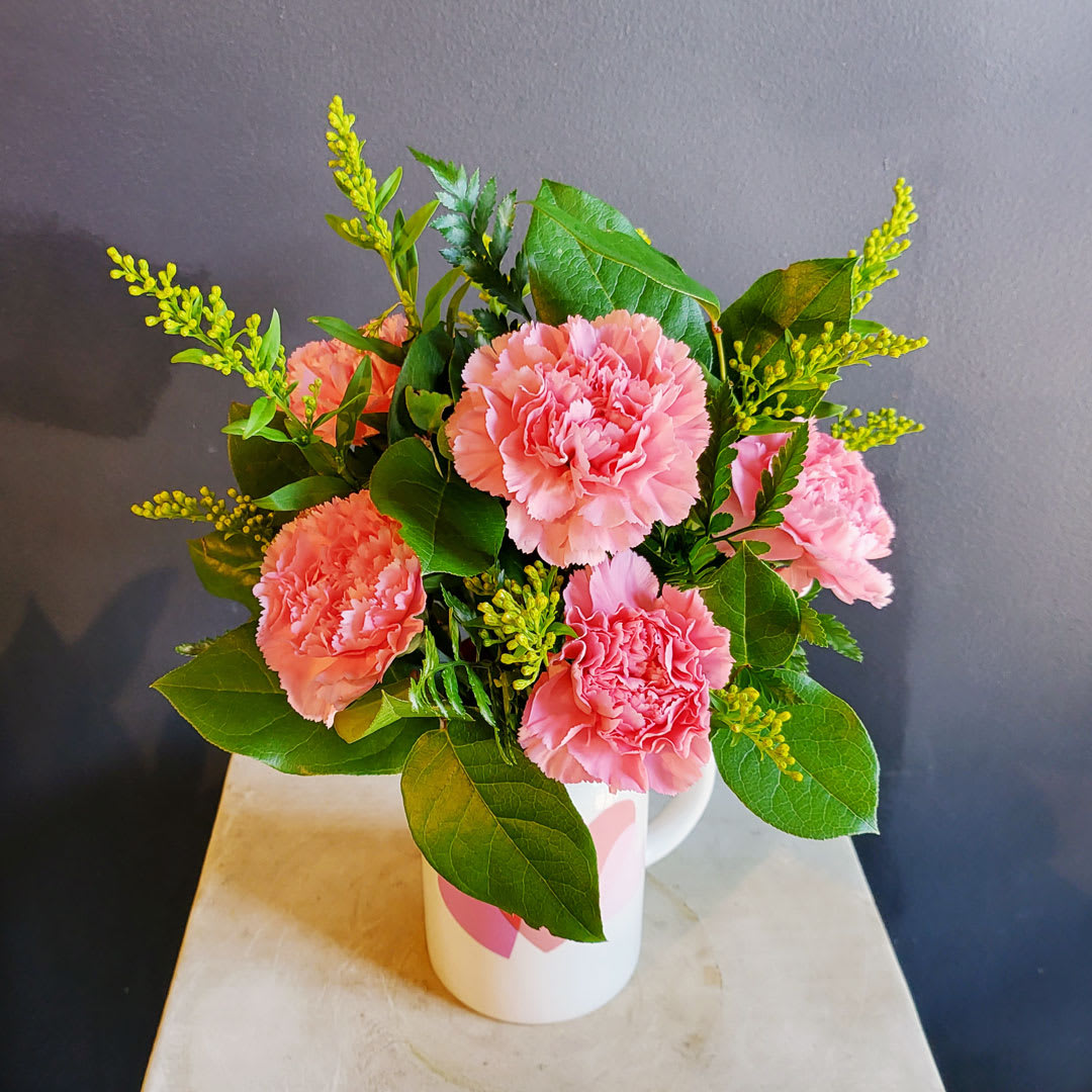 Pink carnations arranged in a white mug with green foliage