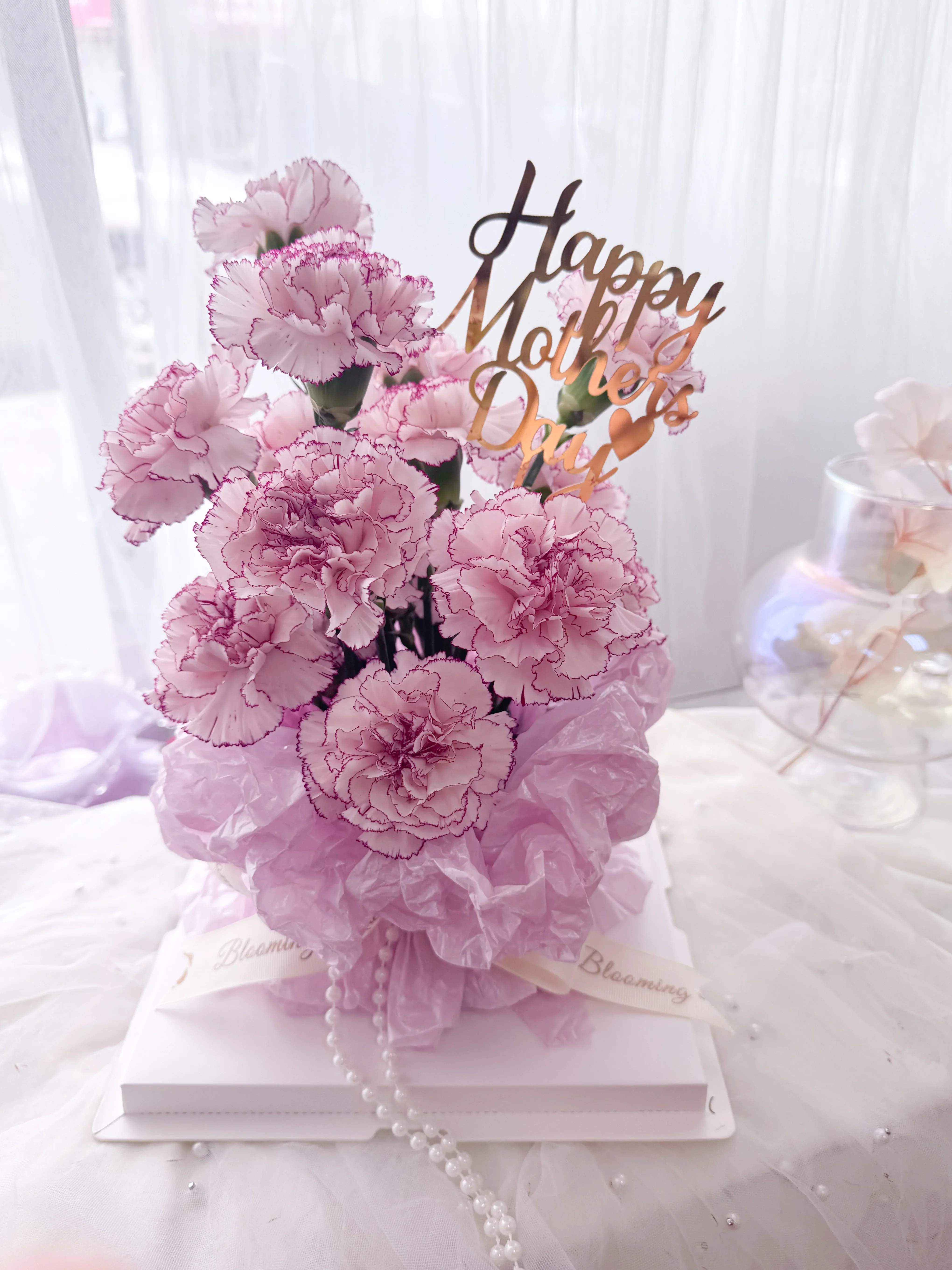 [FRESH FLOWER] Blushing Grace – Mother’s Day Carnation Box - Celebrate her with Blushing Grace, a stunning arrangement featuring 16 stems of novelty violet pink carnations, elegantly displayed in a clear keepsake box. Each bloom is carefully selected for its ruffled beauty and subtle charm, symbolizing a mother’s love in full bloom. Finished with a soft lavender ribbon, blush-toned tissue, pearl accents, and a golden “Happy Mother’s Day” topper, this floral gift is as thoughtful as it is eye-catching—a heartfelt way to say thank you to the most special woman in your life.  *This arrangment already comes with Happy Mother's Day pick. 