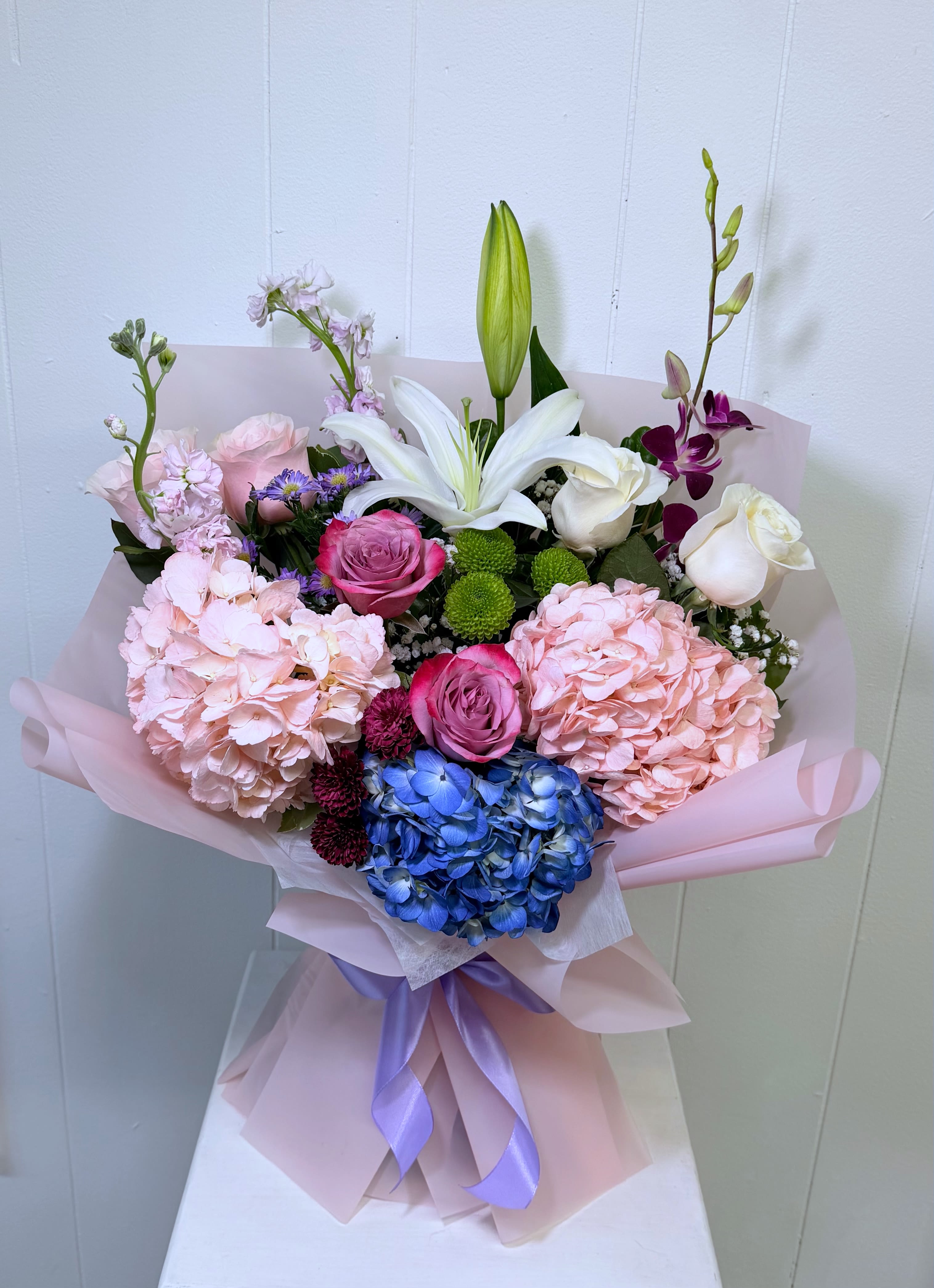   Sweet Elegance Bouquet - A luxurious hand-wrapped bouquet designed with a stunning mix of seasonal blooms including hydrangeas, premium roses, lilies, orchids, green ball chrysanthemums, stock flowers, and fresh seasonal greenery. Wrapped in elegant high-quality paper and finished with a beautiful satin ribbon, this bouquet creates a soft, romantic, and sophisticated presentation.  This bouquet comes with a built-in hydration base, allowing the flowers to stay fresh and beautiful without needing to be placed in a vase immediately. Perfect for gifting, easy to transport, and ready to enjoy exactly as it arrives.  Flower varieties and colors may vary slightly depending on seasonal availability, while always maintaining the same luxurious and elegant style shown in the photo. Perfect for birthdays, Mother’s Day, anniversaries, celebrations, or simply making someone feel truly special.