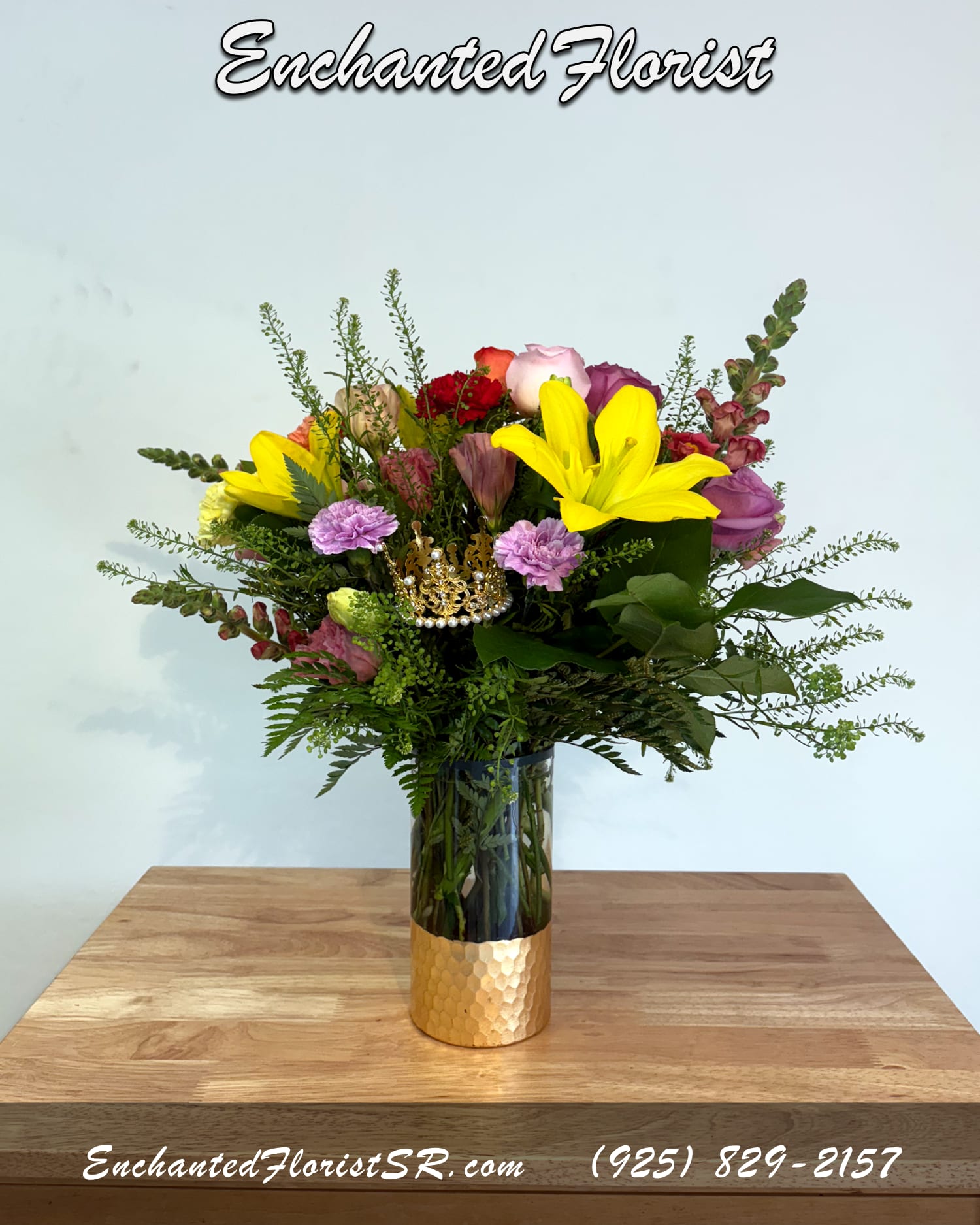 Queen For A Day - Holiday Arrangement - An elegant holiday arrangement showcasing a vibrant multicolored palette. Featuring flowers such as roses, carnations, snapdragons, lisianthus, and lilies. Accented with freshly-cut greens. Presented in a striking cylindrical glass vase that features a golden scale textured base. Accented with a decorative crown nestled within the arrangement offering a whimsical and regal touch.  Perfect for celebrating mothers, grandmothers, and the queens in your life with a gift that feels joyful, heartfelt, and fit for royalty this Mother’s Day.  Orientation: Any-Sided  Arrangement Dimensions (Approximate): Height: 20" Width: 15"  Arrives In: Clear glass cylinder vase with golden scale textured base  Vase Dimensions: Height: 8" Width: 4"