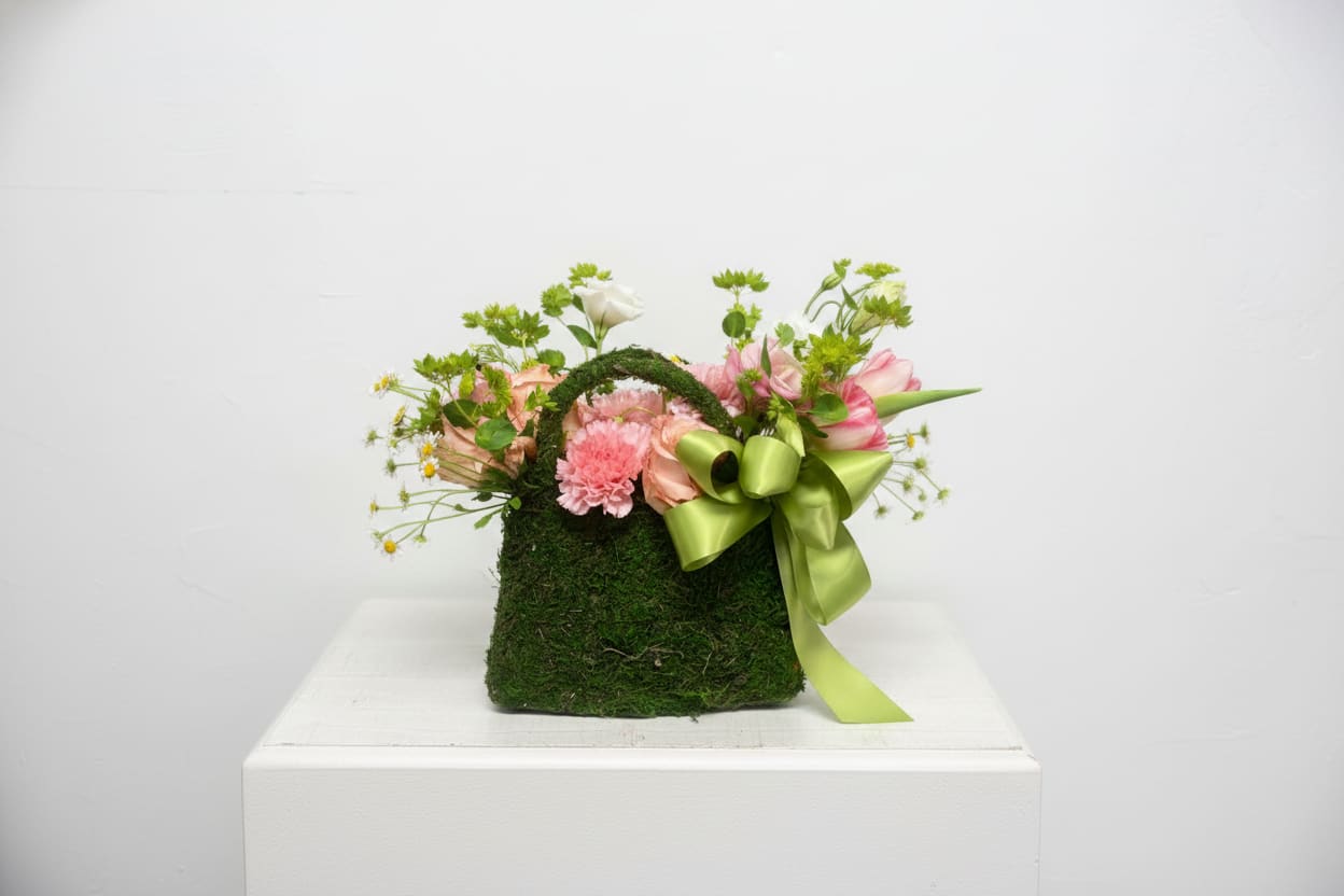     Mossy Meadow Bloom Bag (Seasonal Blooms) - A charming garden-inspired arrangement designed in a moss-covered keepsake bag and filled with soft seasonal blooms in delicate pink, ivory, and fresh green tones. This whimsical design brings together elegant textures and natural beauty for a fresh-from-the-garden feel. Finished with a satin ribbon for an extra touch of sweetness, it’s perfect for birthdays, Mother’s Day, thinking-of-you gifts, or simply brightening someone’s day.  Flowers and accents may vary based on seasonal availability while maintaining the overall color palette and style of the arrangement.