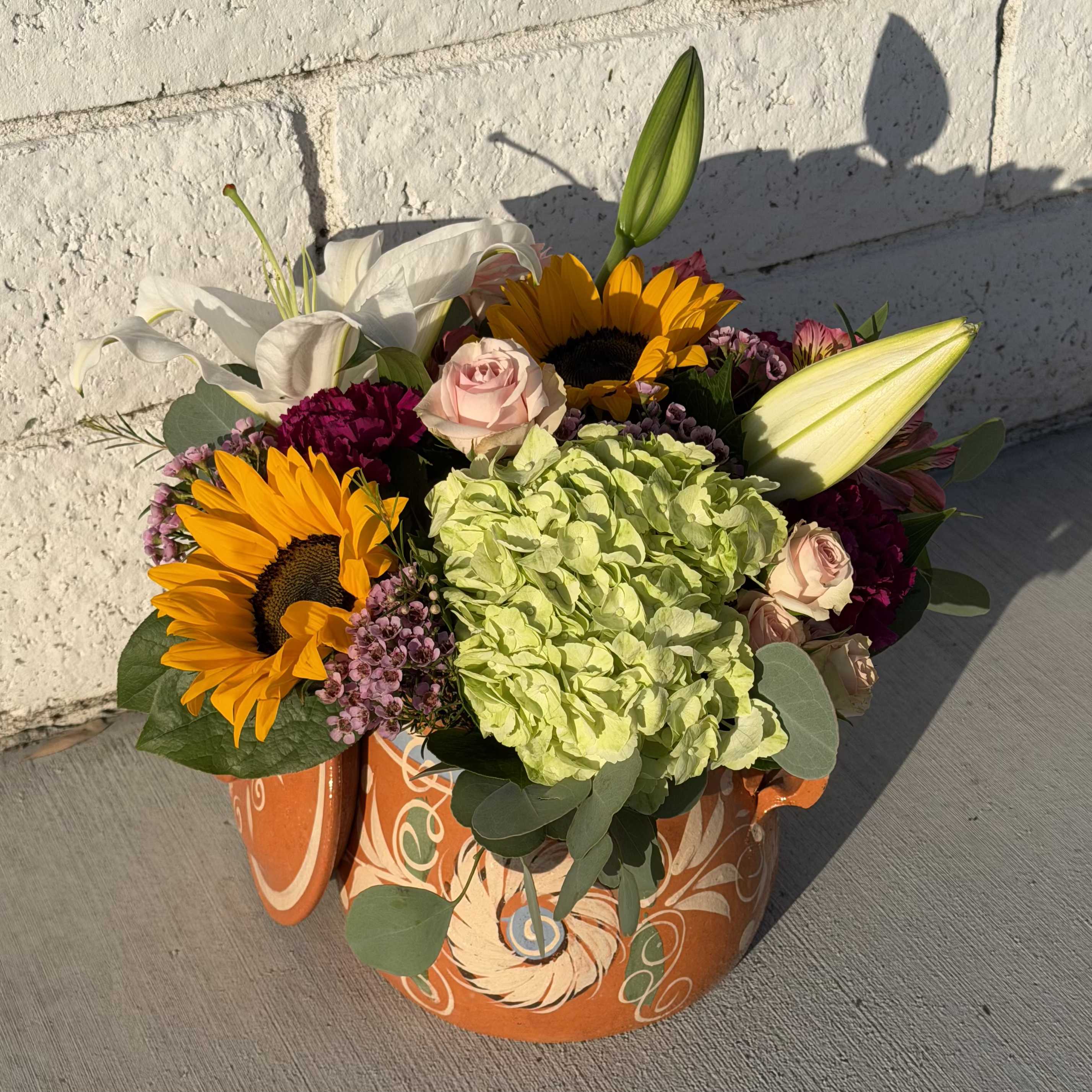 Flores De La Olla Standard  - Bright sunflowers and soft pink lilies make an immediate cheerful impression in this mixed-color flower arrangement for this Mother’s Day.  The arrangement reads as both celebratory and welcoming-the warm yellow of sunflowers and gerbera daisies, the blush and hot-pink roses, and the lime-green hydrangea combine to create a garden-style texture that feels abundant and lively.  This low, rounded design packs blooms tightly for a full, generous look: sunflowers, lilies, roses, hydrangeas, carnations, alstroemeria, gerbera daisies, and purple accent flowers are arranged together for varied color and texture. Expect a mix of yellow, white, pink, purple, and green tones balanced against the patterned orange container.