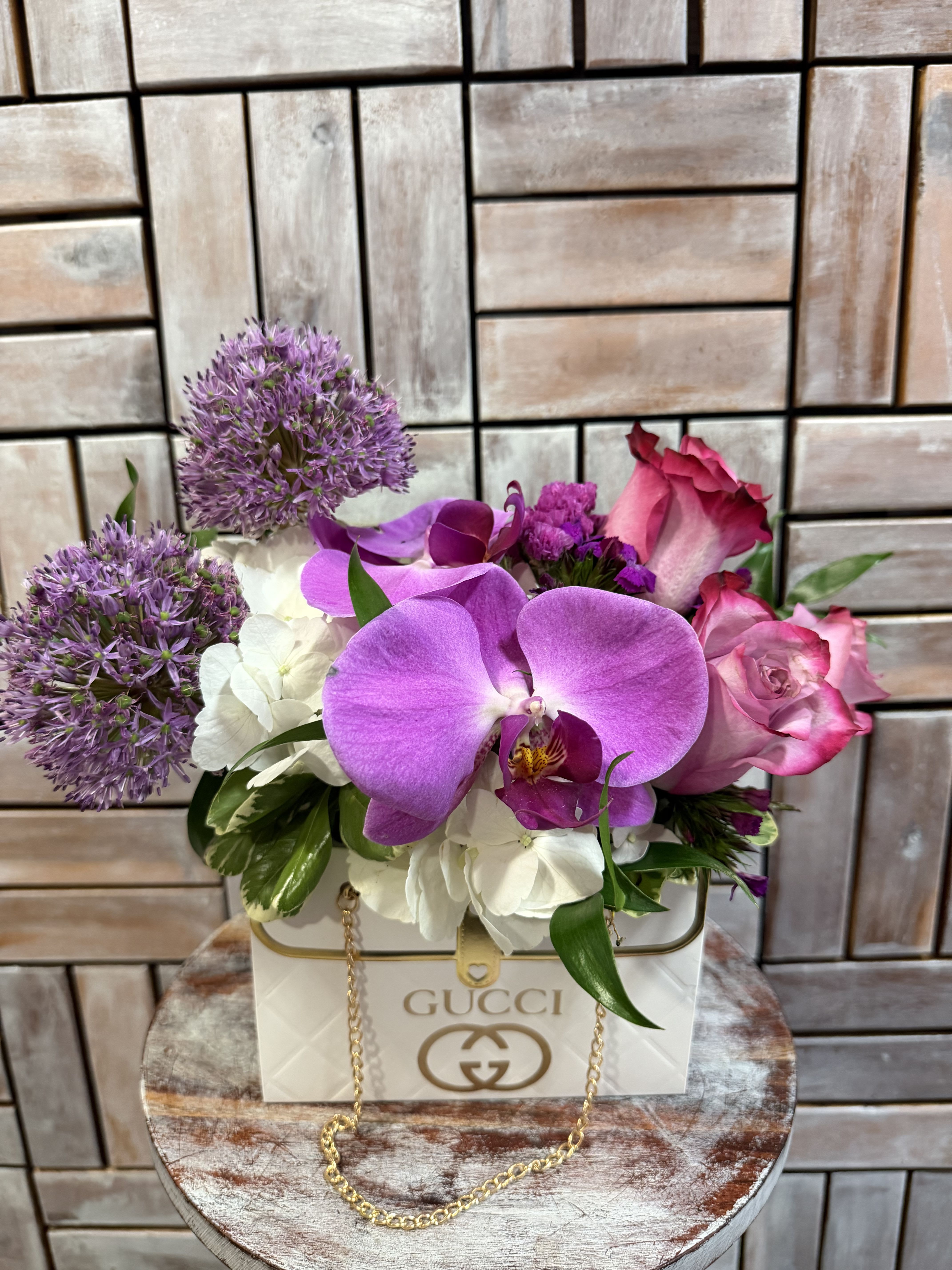 Designer Garden Bloom - A luxurious floral arrangement designed in an elegant, designer-inspired, keepsake handbag container. Featuring vibrant purple orchids, soft pink roses, white hydrangeas, and whimsical allium blooms, this chic design blends modern elegance with high-fashion style. Perfect for birthdays, anniversaries, Mother’s Day, or anyone who loves designer-inspired luxury flowers.