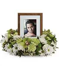 Peaceful Garden Cremation Tribute. - A low, circular sympathy wreath that gently focuses attention on a standing wooden photo frame at its center - perfect for a memorial service or a quiet remembrance display at home. The arrangement reads as a peaceful tribute, the white daisies and chrysanthemums forming a soft, luminous base while green carnations and green cymbidium orchids offer subtle contrast and a sense of calm.  Flowers are densely packed into a continuous ring so the wreath appears full from every angle, creating a neat, finished silhouette around the framed photograph. The palette of white, green, and touches of pale yellow keeps the mood restful and respectful, and the wooden frame adds a warm, personal element ideal for honoring a loved one.  Designed to sit on a table or memorial surface, this arrangement gives the person you're remembering a gentle, dignified focal point that comforts visitors and family. Care is minimal for this funeral-style piece; keep it in a cool, sheltered spot away from direct sun to preserve blooms during the service or while on display. ( Picture Frame Not Included)