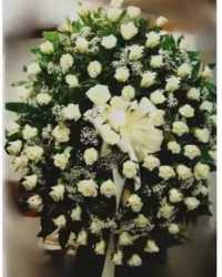 Eternal Light Standing Spray - A full circle of pure white roses and lilies creates a calm, reverent statement perfect for a sympathy arrangement or memorial standing spray. The Eternal Light Standing Spray is designed for funerals and remembrance services-its dense ring of white blooms and gentle trailing ribbon give the person you're honoring a dignified, heartfelt focal tribute.  Tightly clustered white roses form the body of the spray, softened by fine white filler flowers and a prominent cluster of white lilies at the center for texture and a subtle scent. Deep green foliage frames the circular shape for crisp contrast, and a light ribbon trails from the middle to add motion and a final touch of grace. The piece is mounted on a tall metal easel, giving it strong vertical presence at services and viewings.  This classic, formal design reads as respectful and serene-the kind of arrangement that helps mourners feel remembered and comforted. Available in a large, round size appropriate for display on the included easel; please let us know any service details when ordering so we can ensure timely delivery for the memorial.
