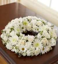 White Cremation Wreath - A low, circular sympathy wreath crafted in pure whites and soft greens offers a calm, reverent tribute for a funeral service or memorial. This sympathy arrangement combines white roses, full-headed chrysanthemums, daisy-style blooms, and carnations to form a continuous ring that reads as one textured, cushion-like expression of comfort-an arrangement meant to make the person remembered feel honored and gently celebrated.  The wreath is densely packed so the petals create a seamless, layered look with subtle green centers for soft contrast, resting low for ease of display on casket lids or memorial tables. Its compact, circular shape and monochromatic palette convey quiet dignity; care instructions are simple-keep cool and hydrated until service time. This is a thoughtful way to communicate sympathy and respect to the family when words alone aren't enough.