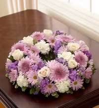 Lavender Cremation Wreath - Soft lavender and pale pink chrysanthemums form a serene, low circular wreath that speaks gently to moments of remembrance and sympathy. This lavender cremation wreath centers an open ring of blooms-lavender daisy-style mums, pale pink chrysanthemums, clusters of white roses and carnations-so the person you're honoring will feel thoughtfully remembered and comforted by the calm, reflective palette.  The arrangement is densely packed around the wreath frame with rounded forms and a blended texture of roses, carnations, chrysanthemums, and daisies in shades of lavender, pink, white, and touches of purple. Its low profile and symmetrical open center make it an elegant display piece for memorial services, a graveside tribute, or a quiet home remembrance on a polished stand or table.  Whether for a funeral, celebration of life, or a private moment of reflection, this wreath offers a comforting, traditional expression of sympathy that reads as tender and respectful. The compact, flat design is ready to lay or display; please contact us for delivery or placement guidance to ensure it is presented exactly as intended.