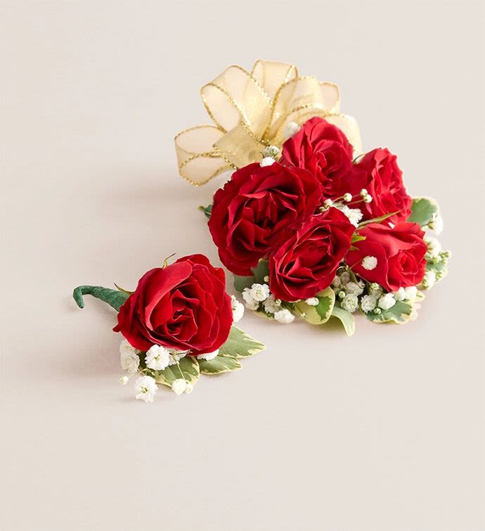 Red Spray Rose Corsage / Boutonniere - A cluster of deep red spray roses makes an immediate romantic statement for weddings, proms, or anniversaries-this matching wrist corsage and boutonniere set is designed to make the person you're honoring feel elegant and noticed. The wrist corsage presents several red blooms gathered tightly and ringed with tiny white filler flowers for a soft, vintage contrast, while the coordinating boutonniere repeats that single-rose look so partners or escorts wear a perfectly paired accent.  Finished with a large, sheer gold ribbon bow and finer gold ribbon accents, the set brings a warm, formal shimmer that reads beautifully in photos and under evening lights. Greens peek through the arrangement to add fresh texture and support; the corsage is multi-bloom and wearable on the wrist, and the boutonniere is a compact single-stem design-ideal for tux lapels or dress jackets. Choose this duo when you want a classic, romantic touch that ties two looks together for a special night.