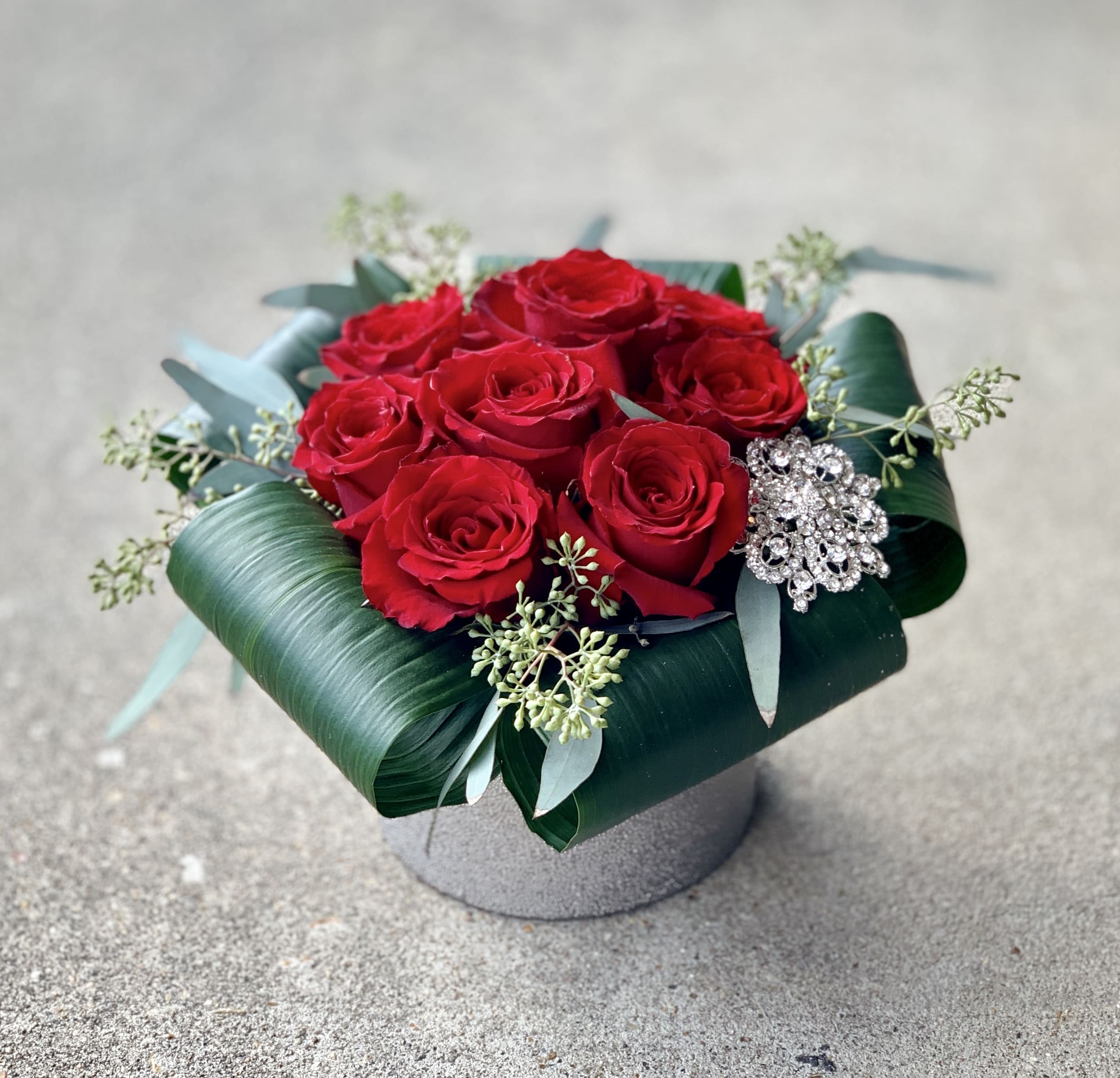 Red roses arranged in a low box with a jeweled brooch accent