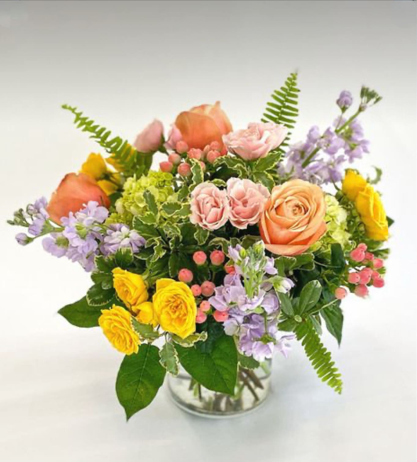 Mixed bouquet of peach and yellow roses, pink spray roses, and lavender blooms in a clear glass vase
