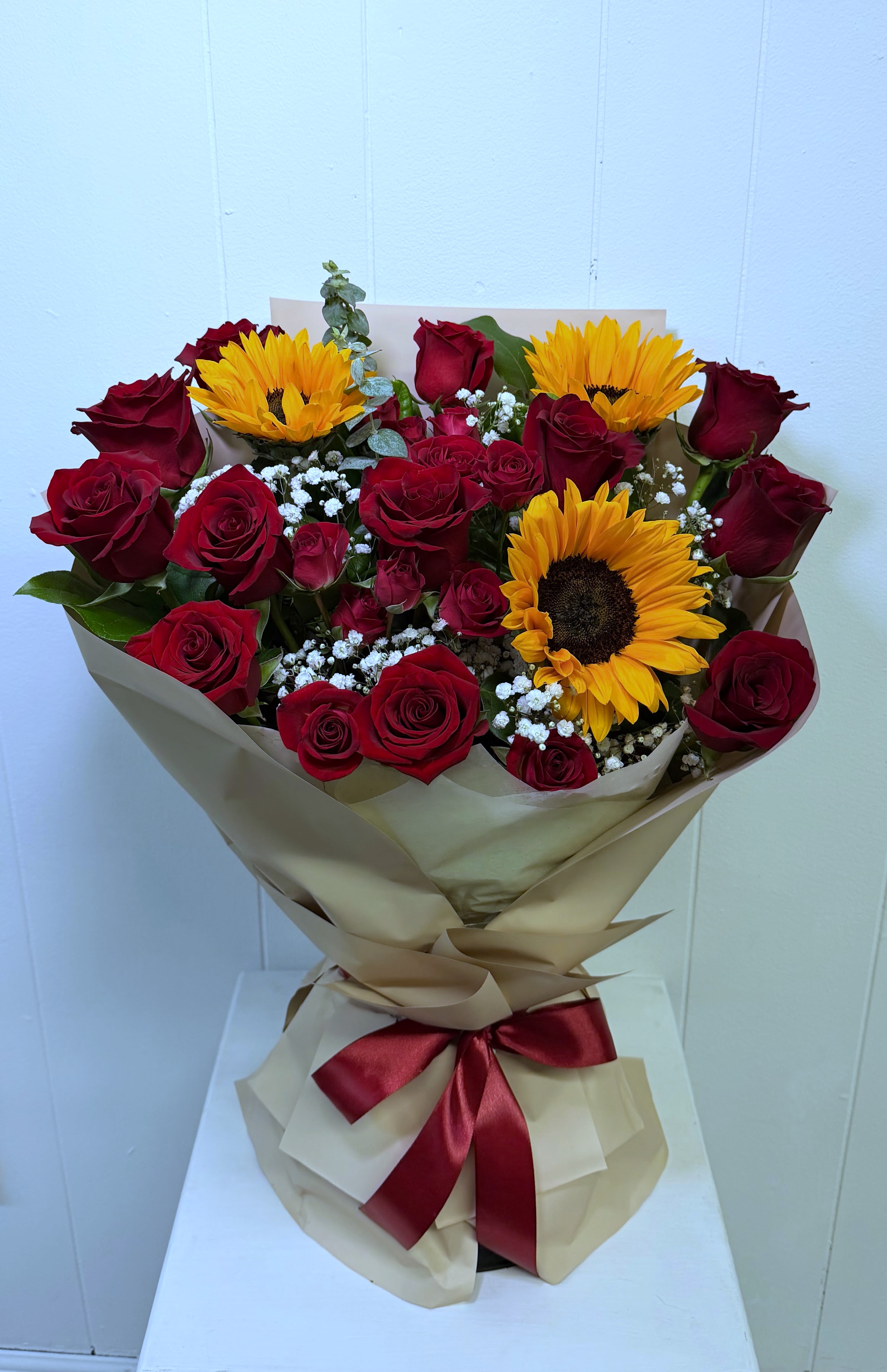 Sunset Love  - A breathtaking hand-wrapped bouquet featuring vibrant sunflowers, premium red roses, fresh greenery, and delicate baby’s breath beautifully arranged in an elegant and romantic style. Wrapped in luxurious high-quality paper and finished with a stunning satin ribbon bow, this bouquet is designed to brighten anyone’s day instantly.  This bouquet comes with a built-in hydration base, allowing the flowers to stay fresh and beautiful without needing to be placed in a vase immediately. Simply keep the bouquet as it arrives and enjoy its elegant presentation and freshness.  A perfect combination of romance and happiness — ideal for birthdays, anniversaries, celebrations, Mother’s Day, or simply making someone feel loved and special.