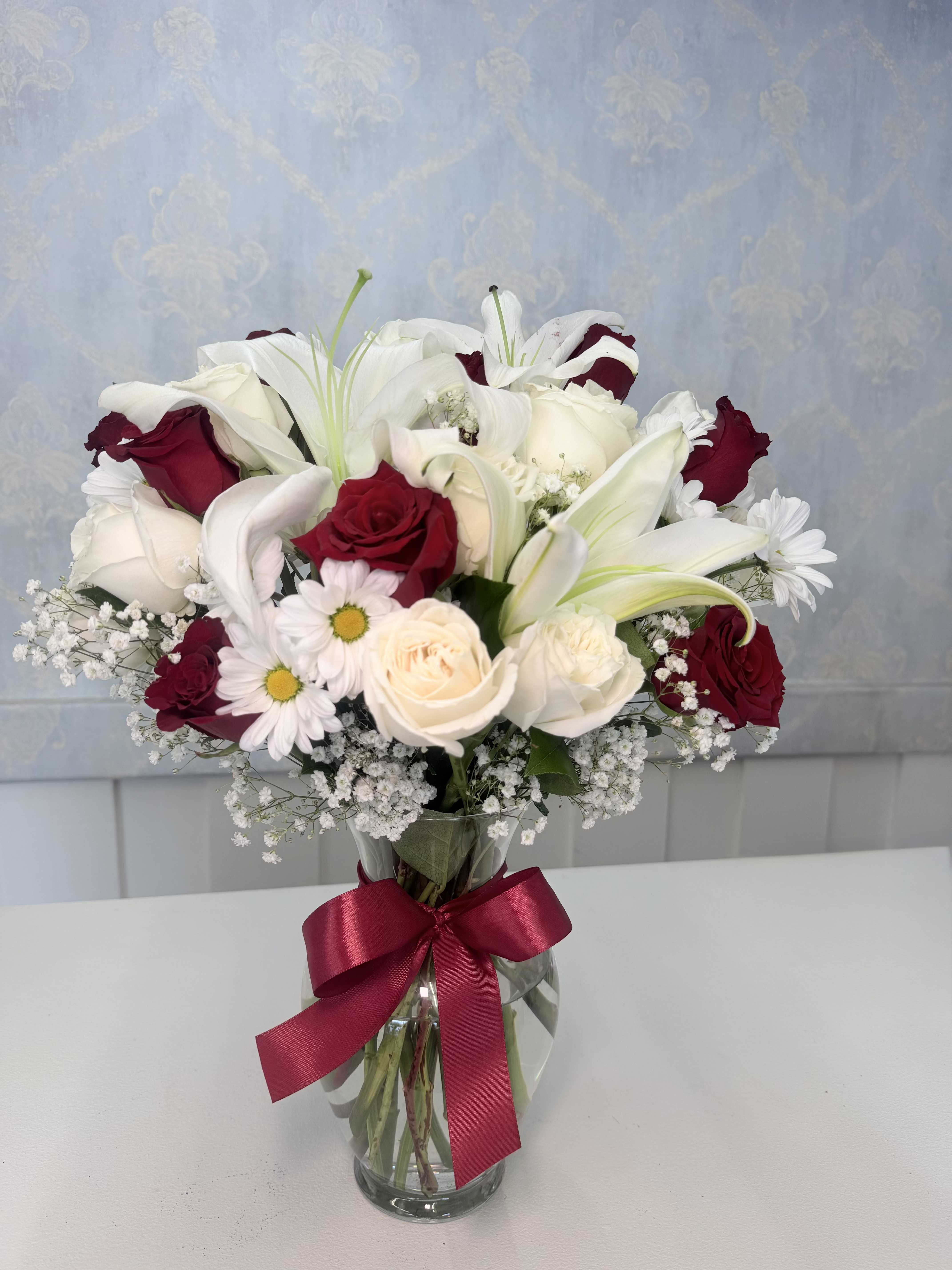   Eternal Romance - Fall in love with the beauty and elegance of our Eternal Romance arrangement  This stunning floral design features a luxurious mix of red and white roses, elegant white lilies, fresh daisies, delicate baby’s breath, and lush greenery arranged beautifully in a clear glass vase.  Finished with a satin red bow, this arrangement is perfect for birthdays, anniversaries, romantic surprises, or any special occasion where you want to make someone feel truly loved.  A timeless arrangement filled with elegance, freshness, and romance that will brighten any room instantly.