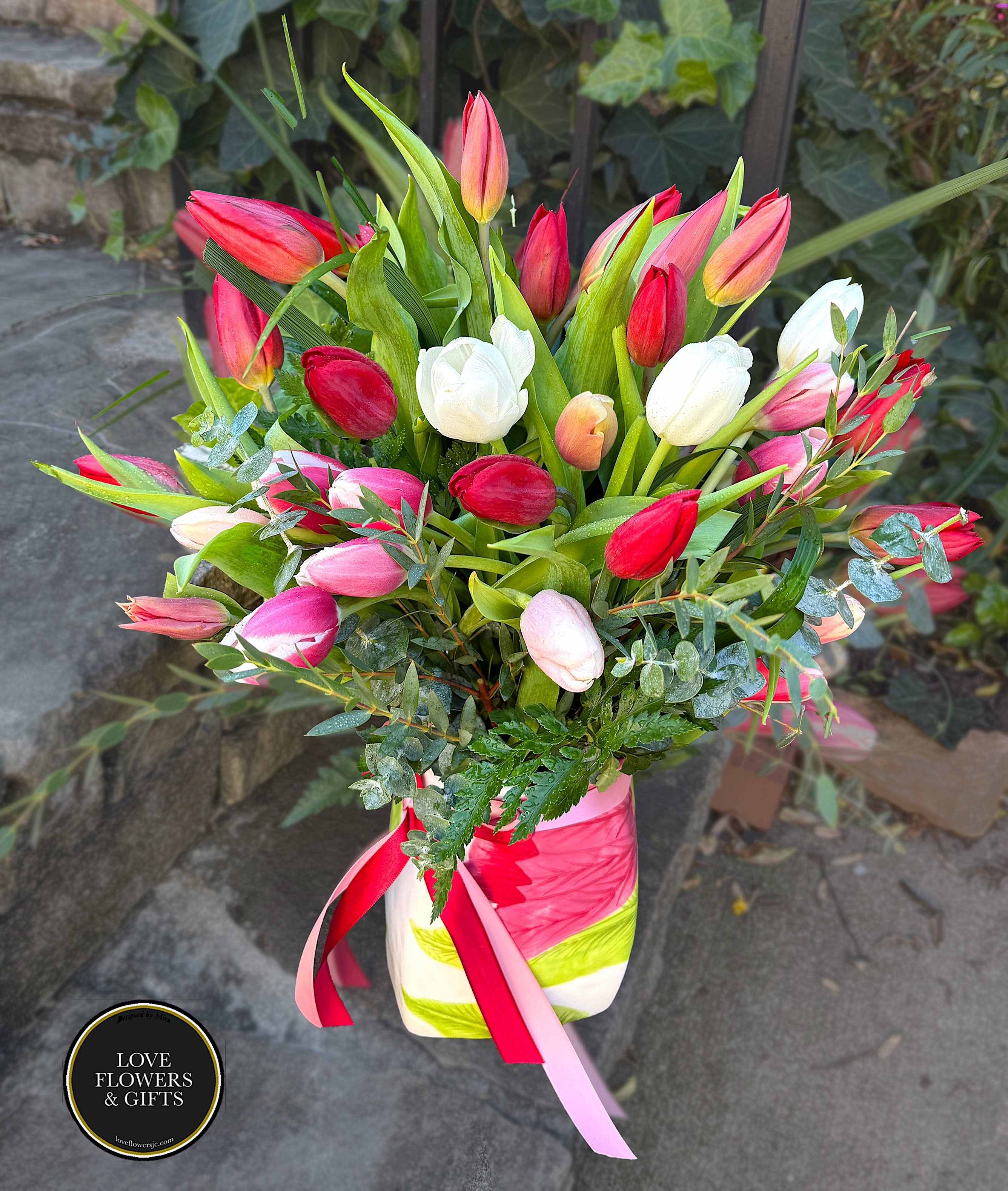 Tall arrangement of red, pink, white, and peach tulips in a striped vase with ribbons