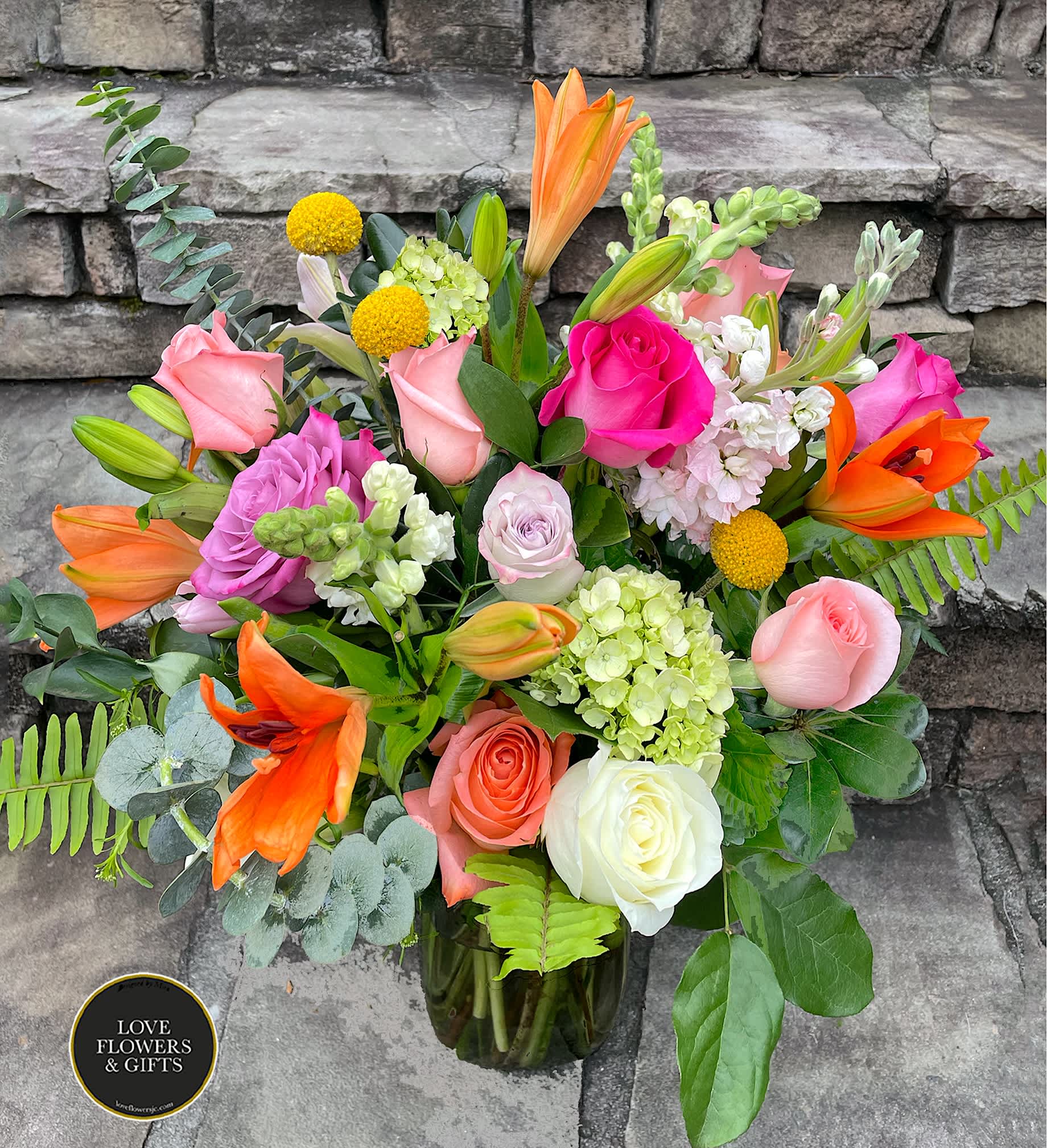 Colorful arrangement of pink roses, orange lilies, and mixed blooms in a clear glass vase.