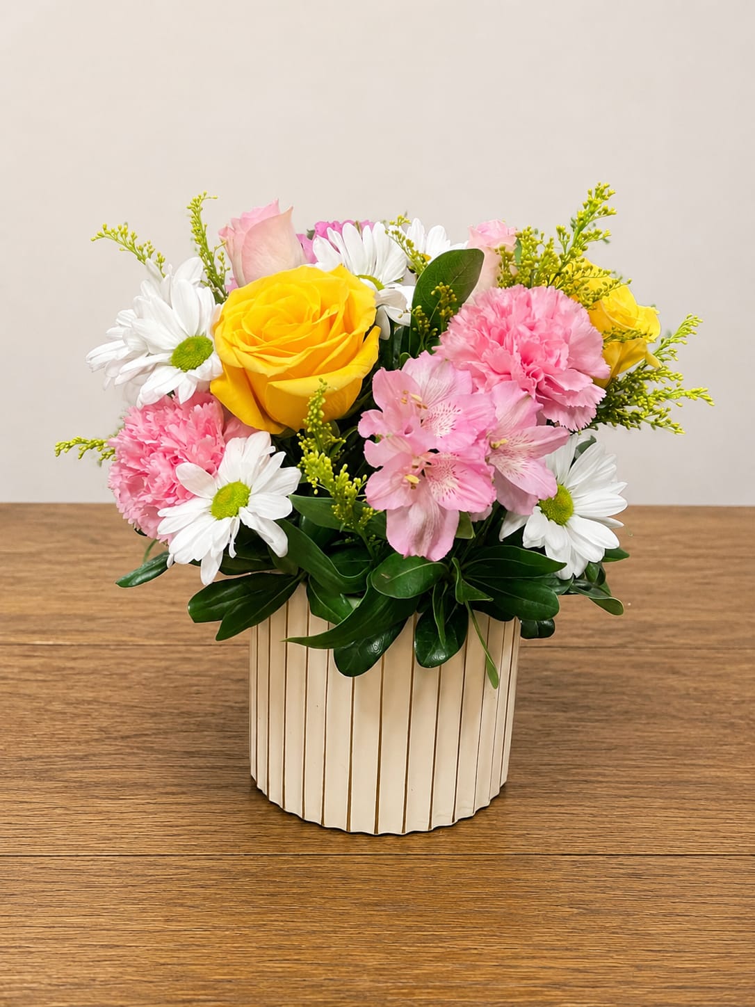 Watercolor - A charming blend of soft romance and fresh elegance, this arrangement features creamy yellow roses, blush pink blooms, and delicate white daisies nestled in lush greenery designed in a modern ribbed ceramic vase.