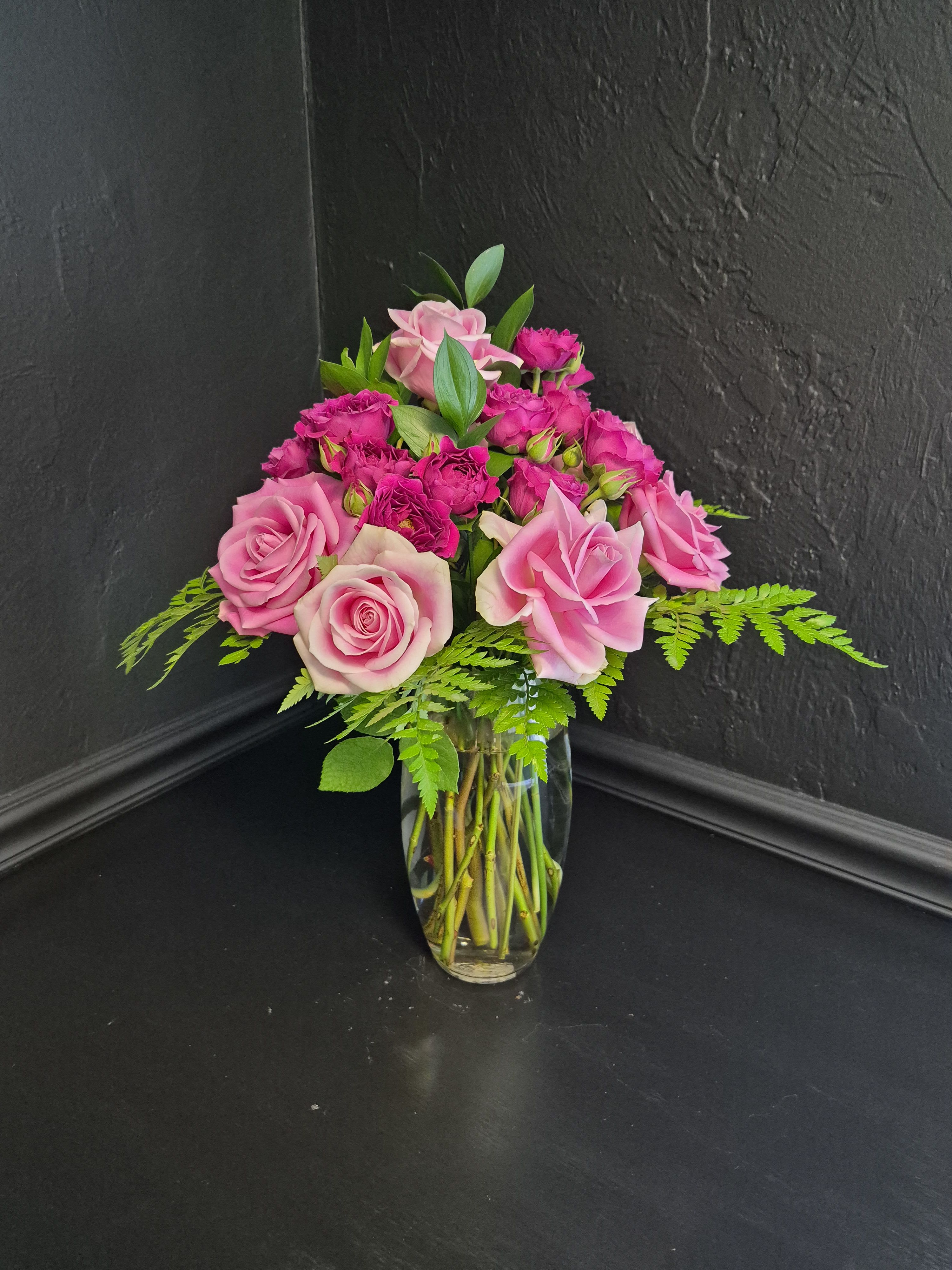 Rosé All Day - A lush cluster of blush pink roses and vibrant magenta spray roses creates an instantly romantic arrangement that says “you’re on my mind” without saying a word. The rounded, garden-style shape and rich pink palette make this bouquet perfect for birthdays, anniversaries, thoughtful surprises, or simply brightening someone’s day.  Densely layered blooms are arranged in a clear glass vase with airy fern greenery and fresh foliage that add texture and natural contrast. The combination of full roses and delicate spray roses creates beautiful depth, softness, and a lush, elegant presence, while the low rounded silhouette makes it ideal for dining tables, desks, or bedside spaces.  Designed to feel both luxurious and heartfelt, this arrangement arrives beautifully styled and ready to enjoy. To keep blooms looking fresh, change the water and trim stems every few days.