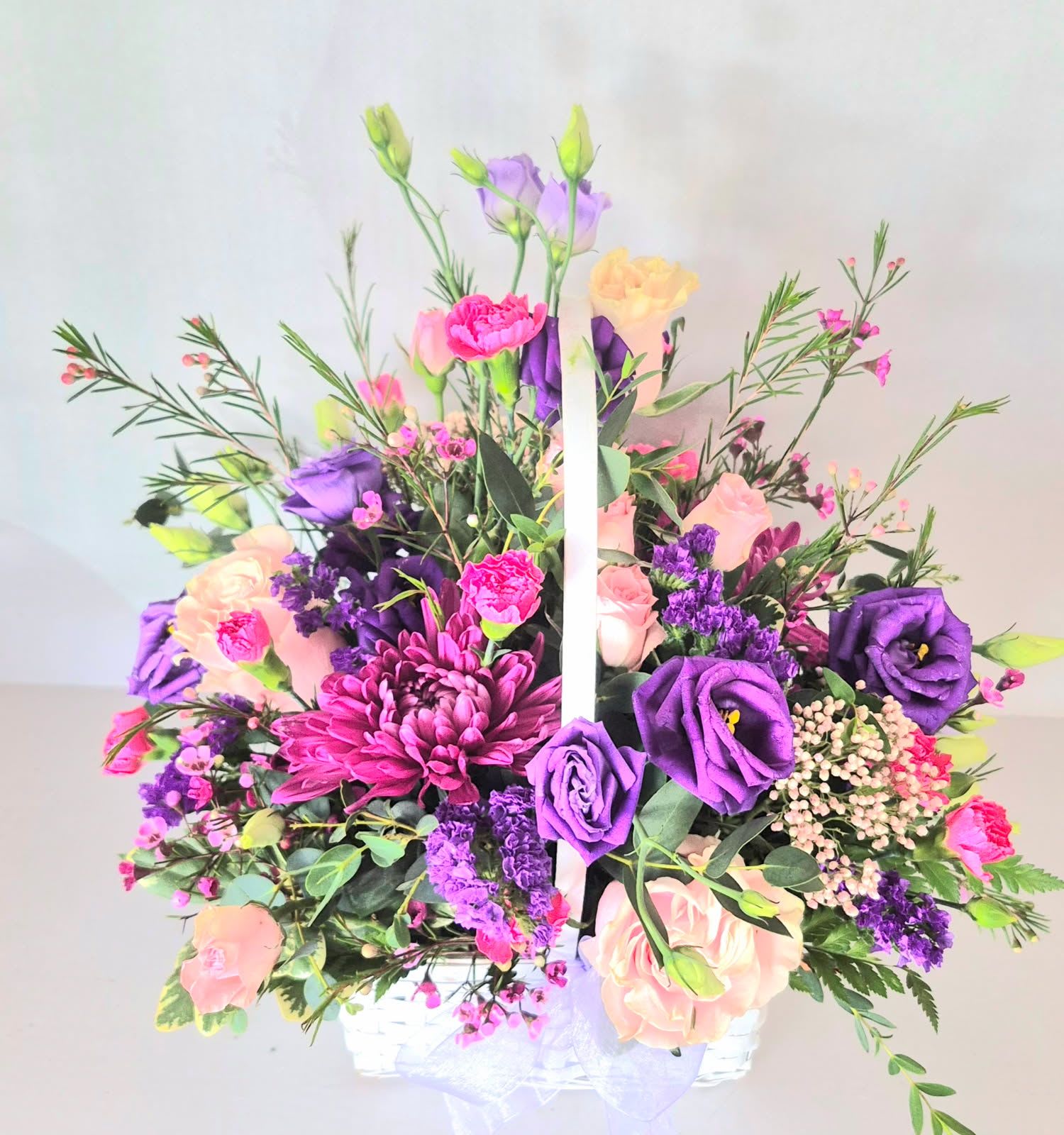 Basket arrangement of purple, pink, and peach flowers with a white handle