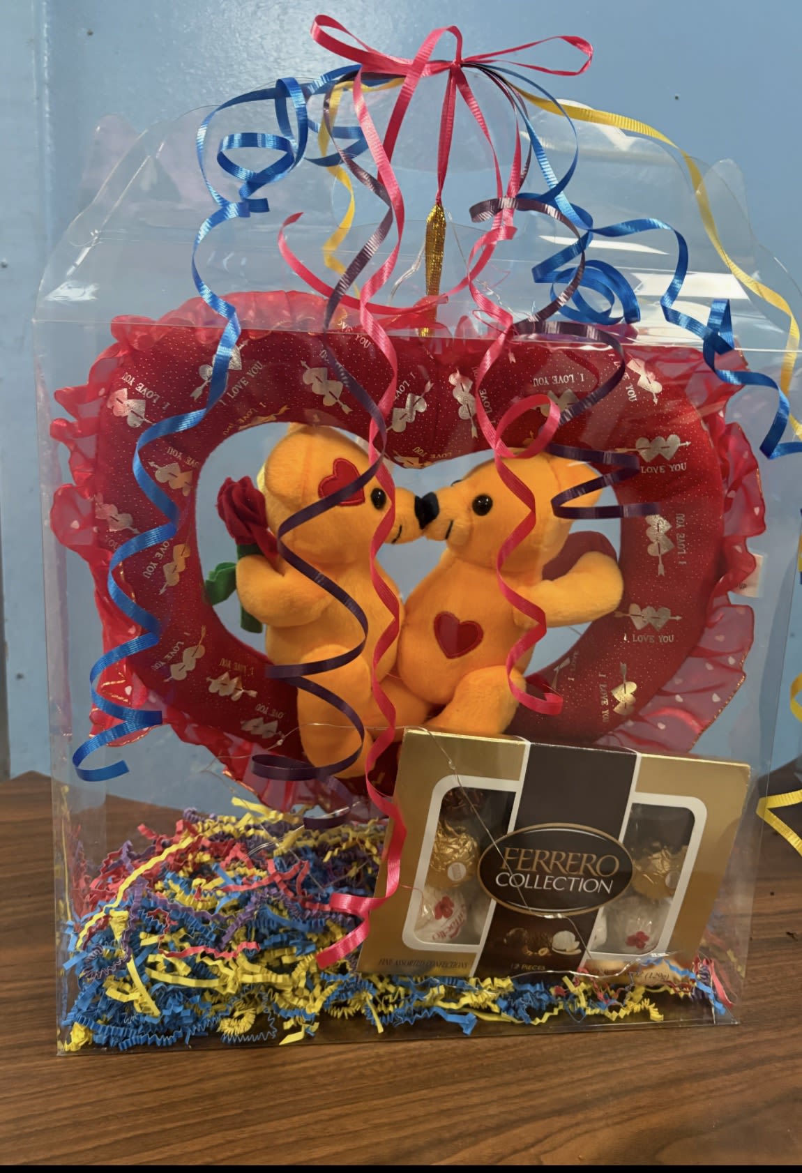 Hey Boo  - Playful, romantic, and impossibly giftable - this Valentine's Day or anniversary flower arrangement comes as a ready-to-give keepsake inside a clear plastic gift box, perfect for surprising someone special. The arrangement frames two small yellow-orange plush teddy bears against a red heart-shaped backdrop printed with love text, sending an unmistakable message of affection the moment the recipient opens it.  Bright curled ribbons in blue, pink, yellow, and purple tumble around the display for a burst of color, while a Ferrero Collection chocolate box at the base adds a sweet complement to the keepsake vibe. The presentation uses a compact, box-style format with decorative ribbon curls and a red heart-shaped backing to create a playful, romantic tableau that feels both celebratory and intimate.  Ideal for a partner, sweetheart, or someone you want to make feel cherished, this boxed arrangement is the kind of gift that delights at first sight and keeps giving - cuddly plush, chocolate treats, and a heart-filled display they can set out as a reminder. Ready-to-ship in its clear gift box, it's an effortless, thoughtful choice when you want them to feel truly seen.