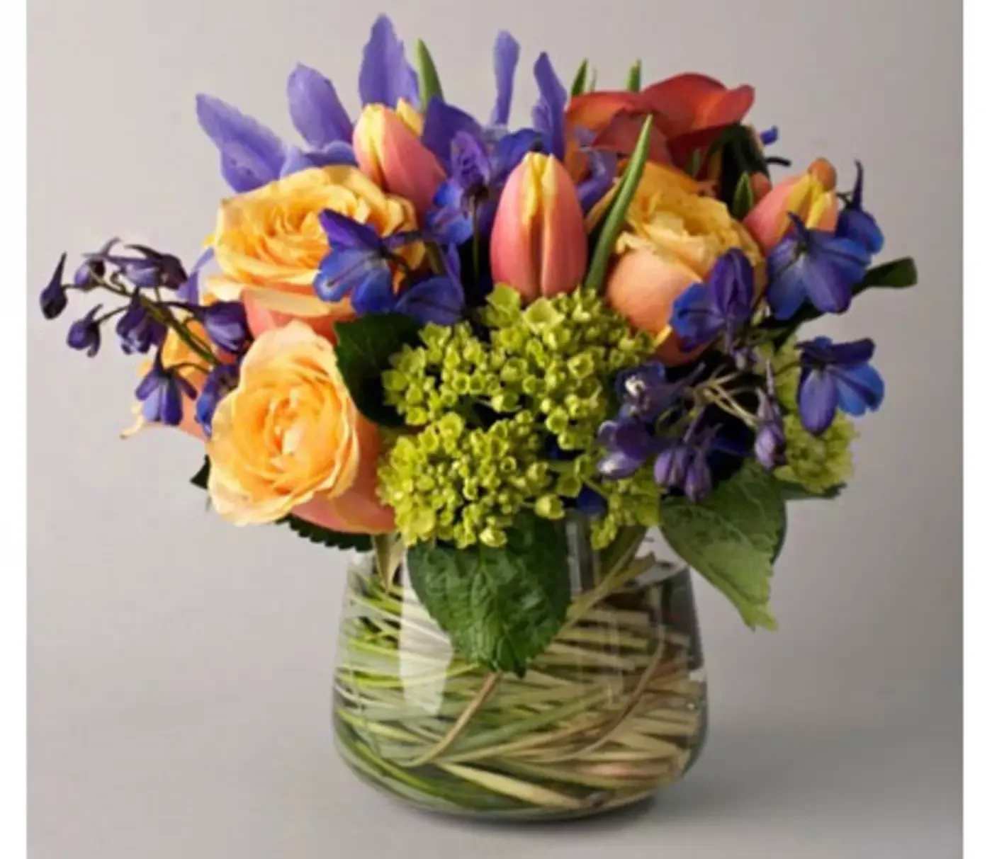 Mixed bouquet of orange roses, pink tulips, and purple flowers in a glass vase