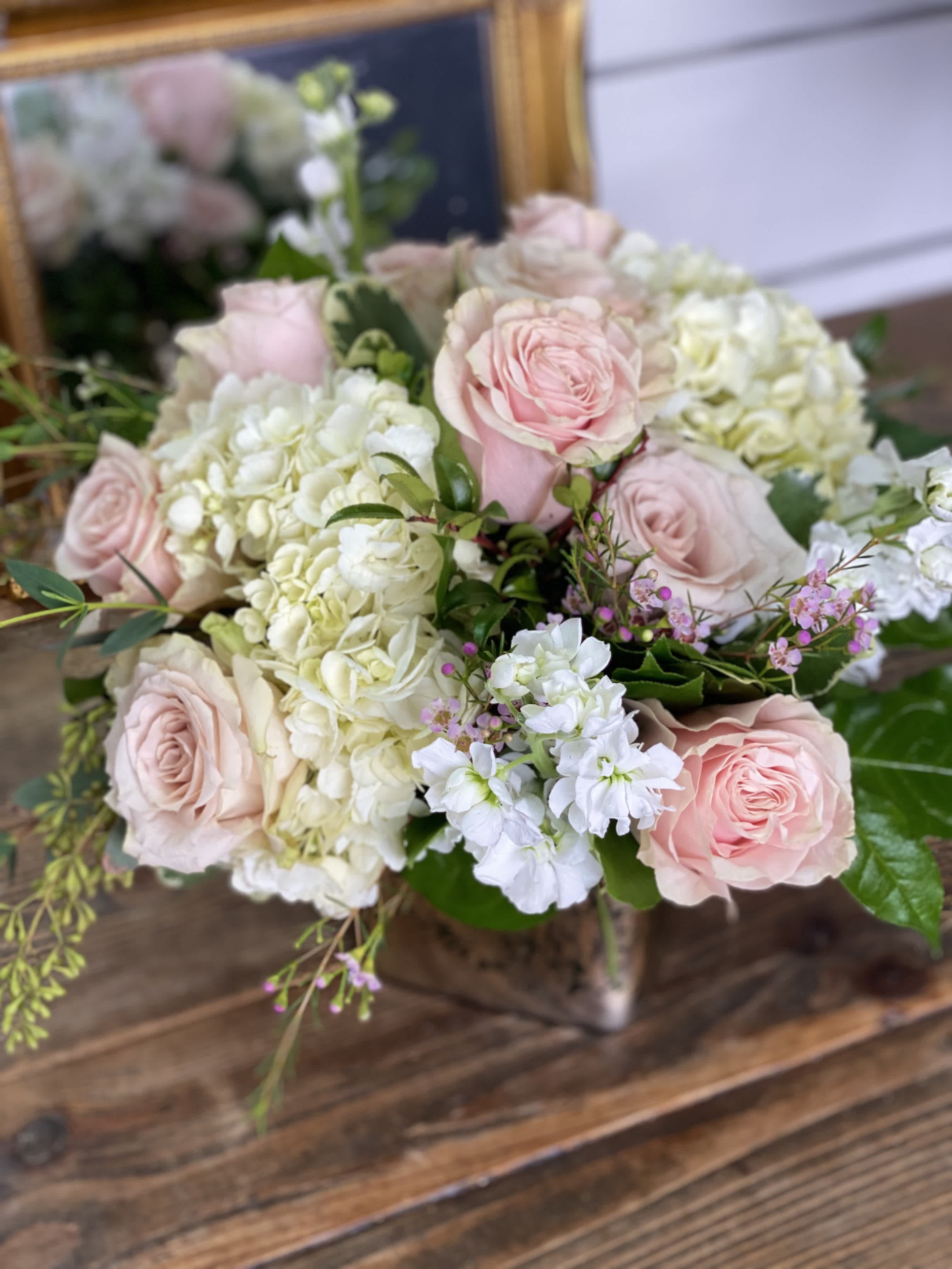 Low arrangement of blush pink roses, white hydrangeas, and white stock in a metallic cube vase on a wooden table.