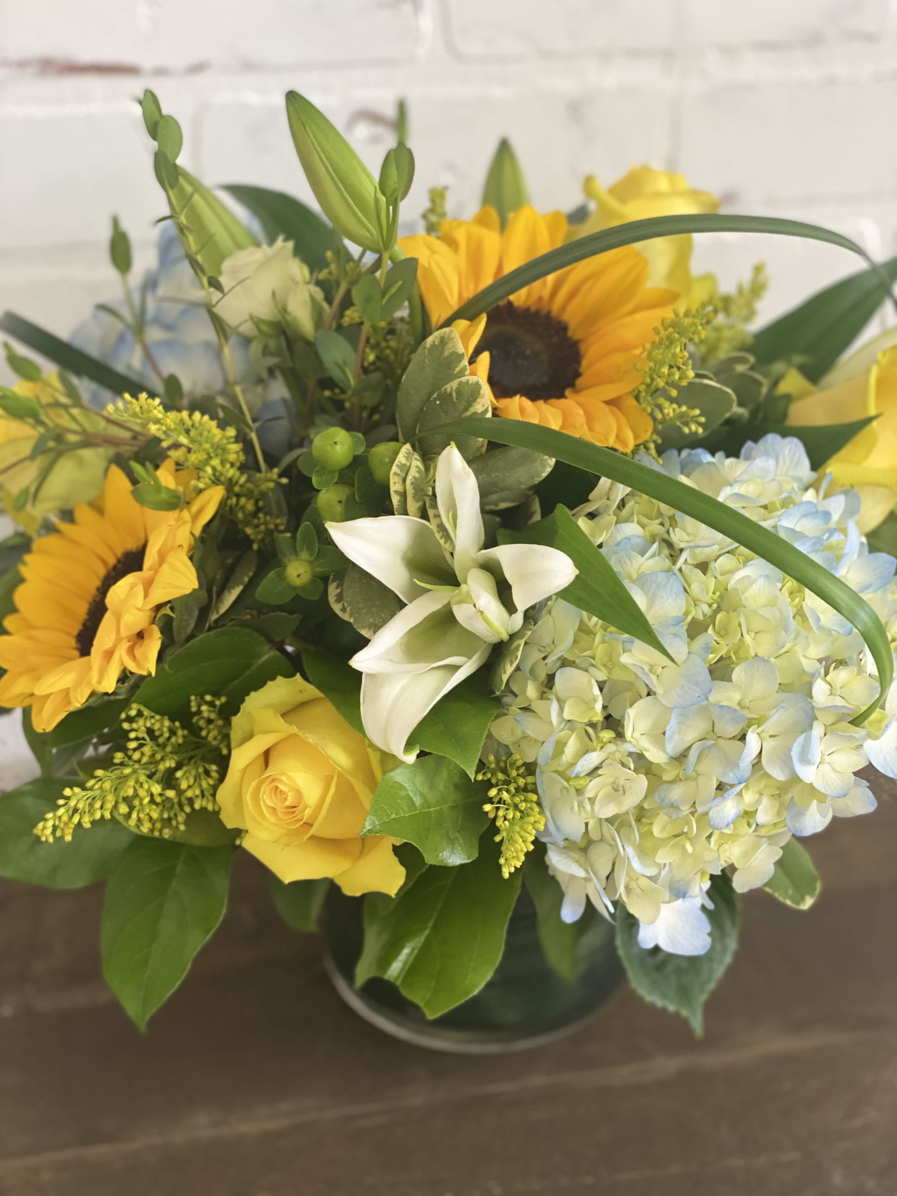 Bouquet of yellow sunflowers, roses, and pale blue hydrangeas in a glass vase