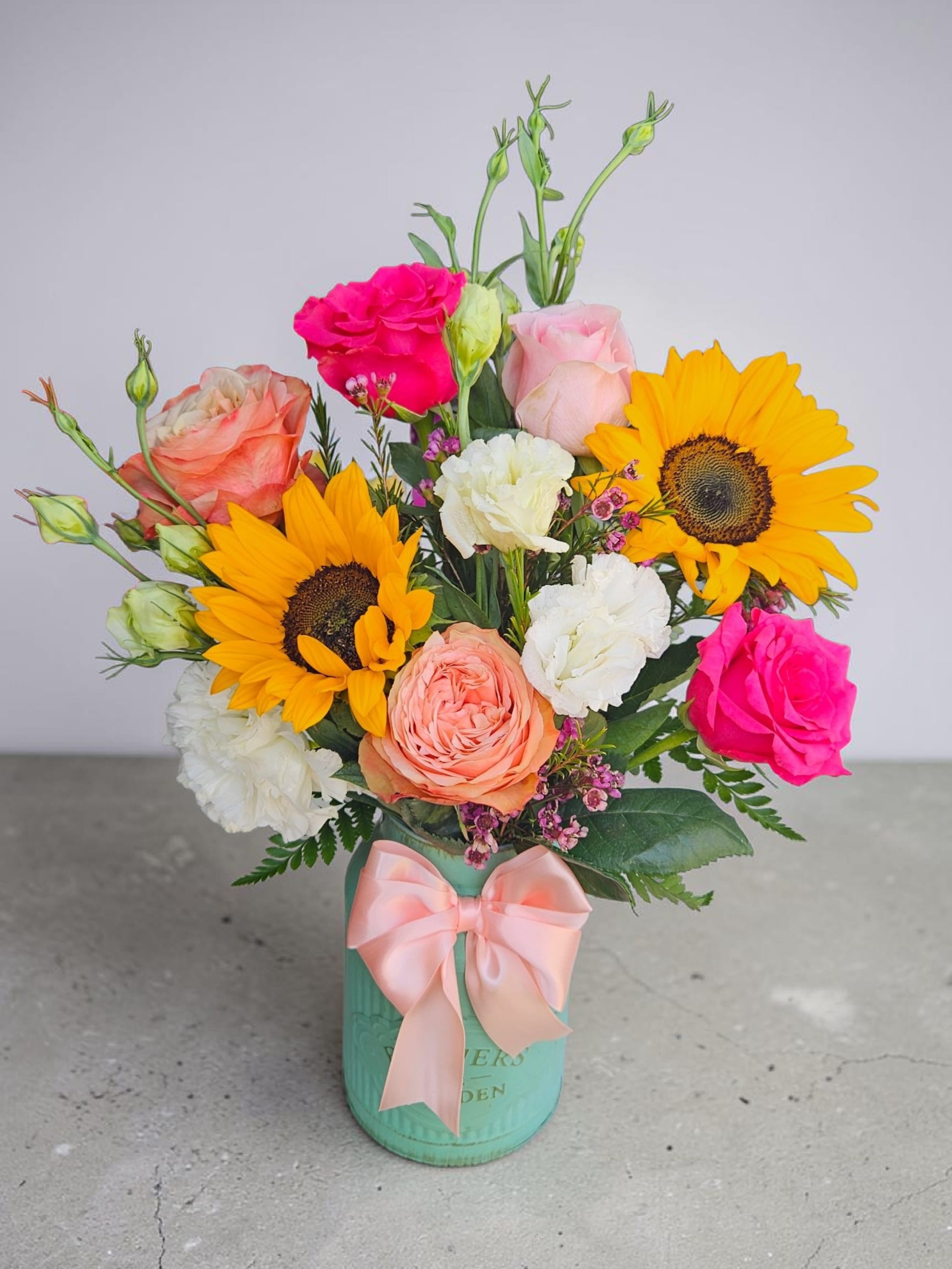 Garden love  - Bright yellow sunflowers burst from a compact, rounded flower arrangement that feels like a pocket of sunshine-perfect as a birthday bouquet, a thank-you gift, or a just-because pick-me-up. Hot pink, blush, peach, and white roses and ruffled carnations weave through the sunflowers to create a layered, garden-style composition that reads lively and romantic; the overall impression is cheerful and celebratory, ideal for making the person you're thinking of feel truly seen.  The mix of rose forms and textured carnations gives the bouquet depth and a soft, tactile rhythm while the sunflowers provide bold contrast and warmth. Presented in a teal/turquoise hard container-choose between a metal vase or a matching hatbox-each option is finished with a pale pink satin ribbon for a polished, gift-ready look. The arrangement's rounded, compact size makes it a versatile centerpiece or bedside surprise; it's designed to arrive fresh and ready to brighten someone's day.