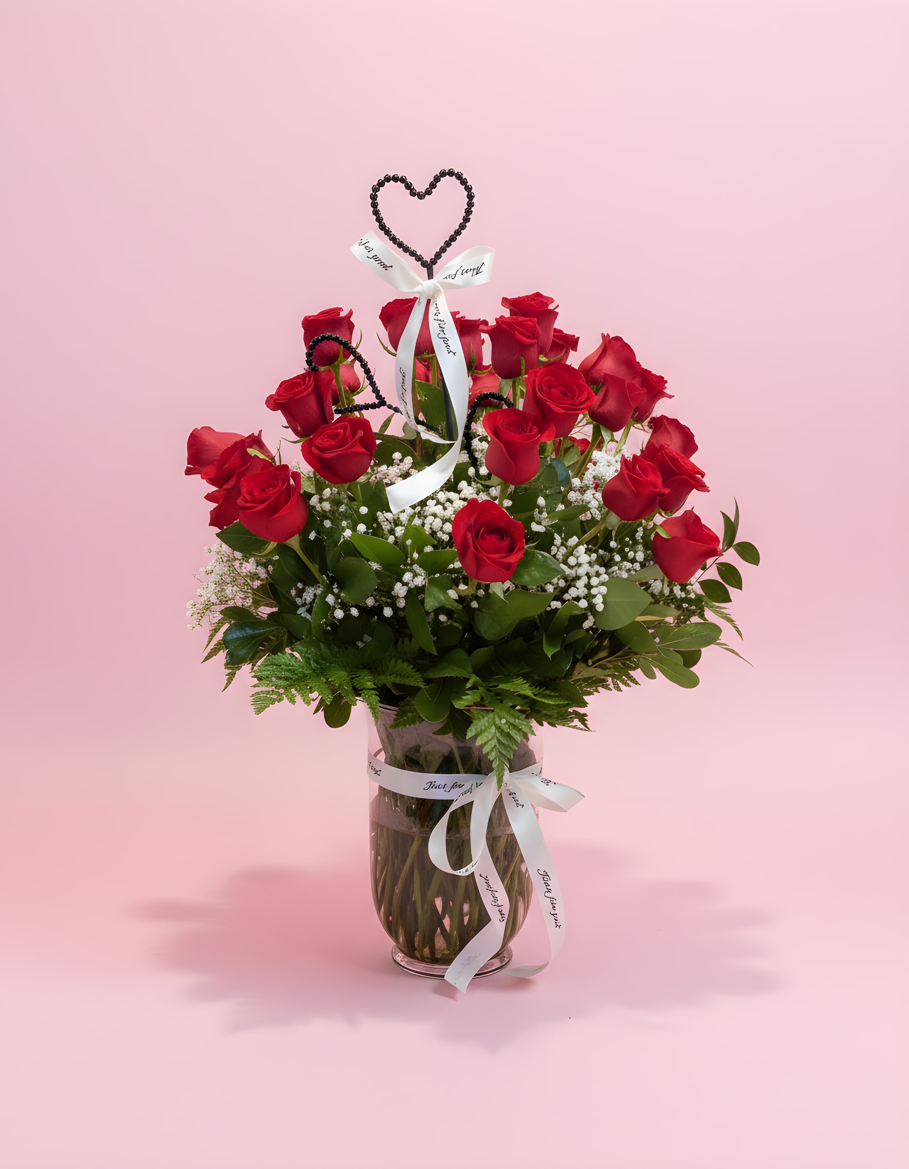 SHOW YOUR LOVE 3 DOZEN ROSES - A towering cluster of three dozen red roses makes an immediate romantic statement-perfect as a Valentine's Day or anniversary bouquet that says "I love you" without words. The tall clear glass vase elevates the blooms so the person you're celebrating feels truly admired; the dense red roses, crisp white accents, and lush greenery create classic, heart-stopping contrast for a meaningful declaration.  The arrangement pairs deep red roses with touches of white baby's breath and abundant green foliage, finished with a white ribbon and a heart-shaped topper for an extra romantic flourish. Presented in a clear glass vase, the design reads as a formal, luxurious display that fills a room and draws the eye. The black beaded heart topper adds texture and a striking focal detail to the overall look.  This is the kind of bouquet that makes someone feel cherished on big romantic milestones-anniversary dinners, Valentine's surprises, or to celebrate a significant "I love you." Arrives ready to display in its tall vase; trim stems and fresh water as needed to prolong vase life.