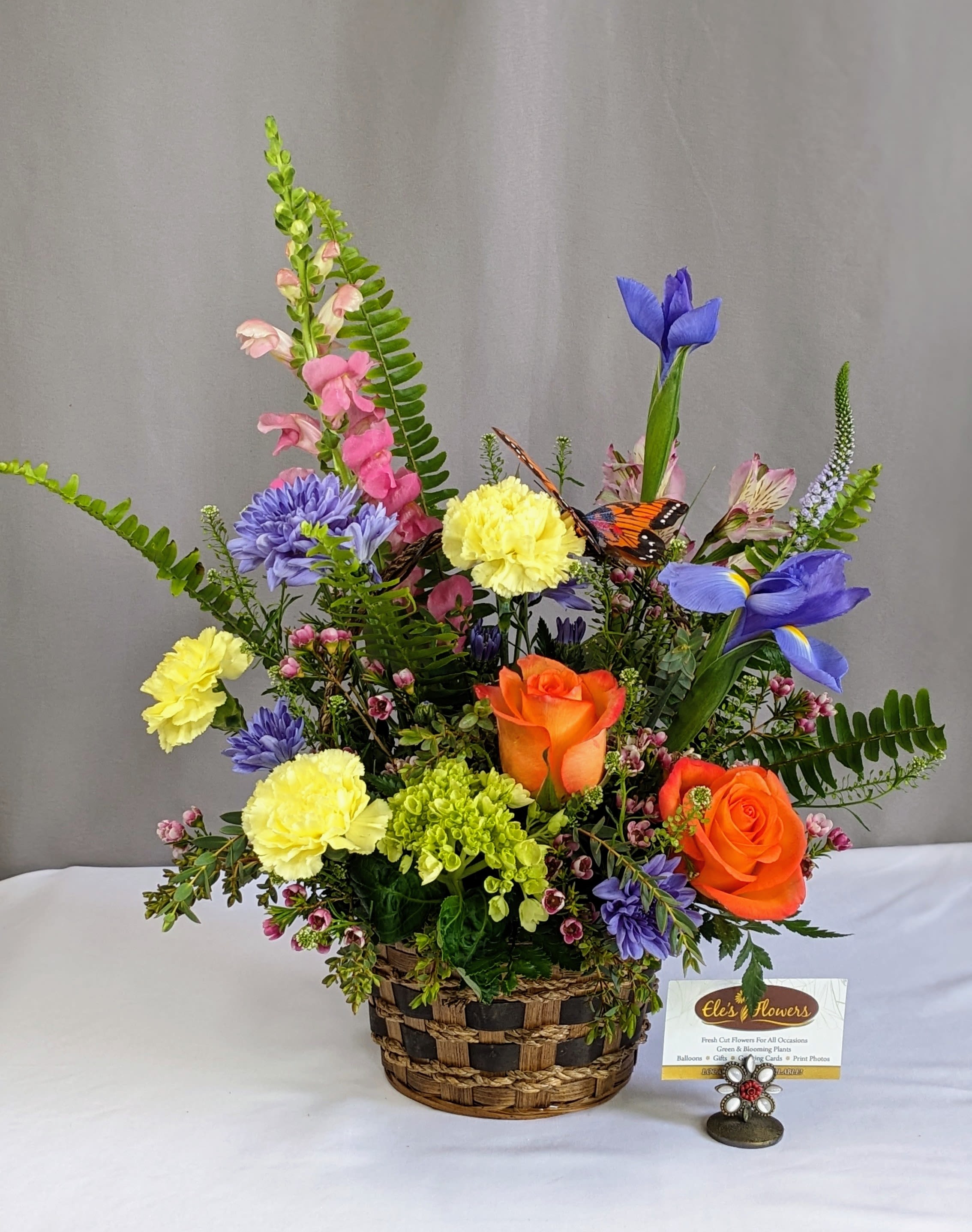 Spring Flutter Basket - This cheerful spring arrangement bursts with fresh, vibrant color and natural movement, creating a lively garden-inspired look. Designed in a woven basket, it features a playful mix of soft pastels and bold pops of color, accented by airy greenery and a delicate butterfly detail for added charm. The varying heights and textures give it a whimsical, just-picked feel—perfect for brightening a room, celebrating the season, or sending a joyful surprise.  Approximate dimensions: 14–16” H x 12–14” W