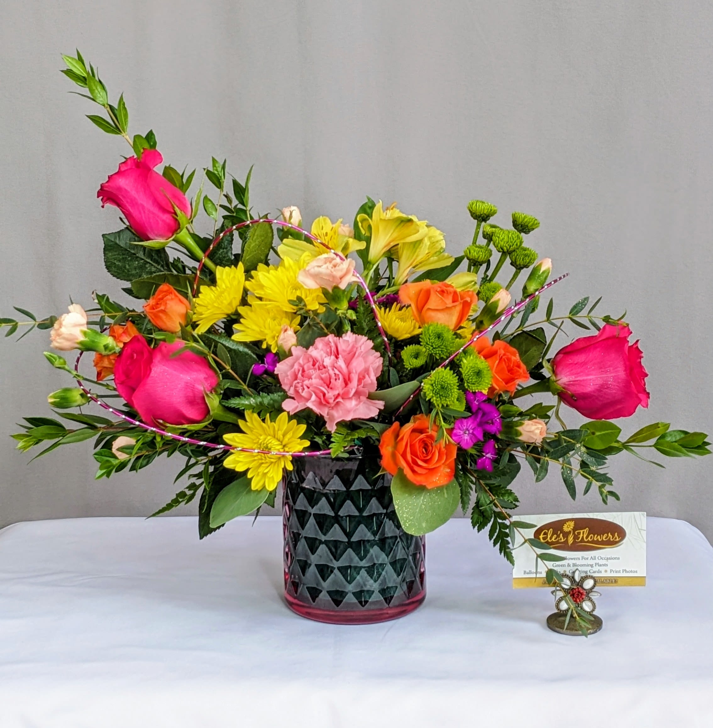 Bright mixed arrangement with pink roses, orange roses, yellow mums, and green button mums in a dark patterned glass vase