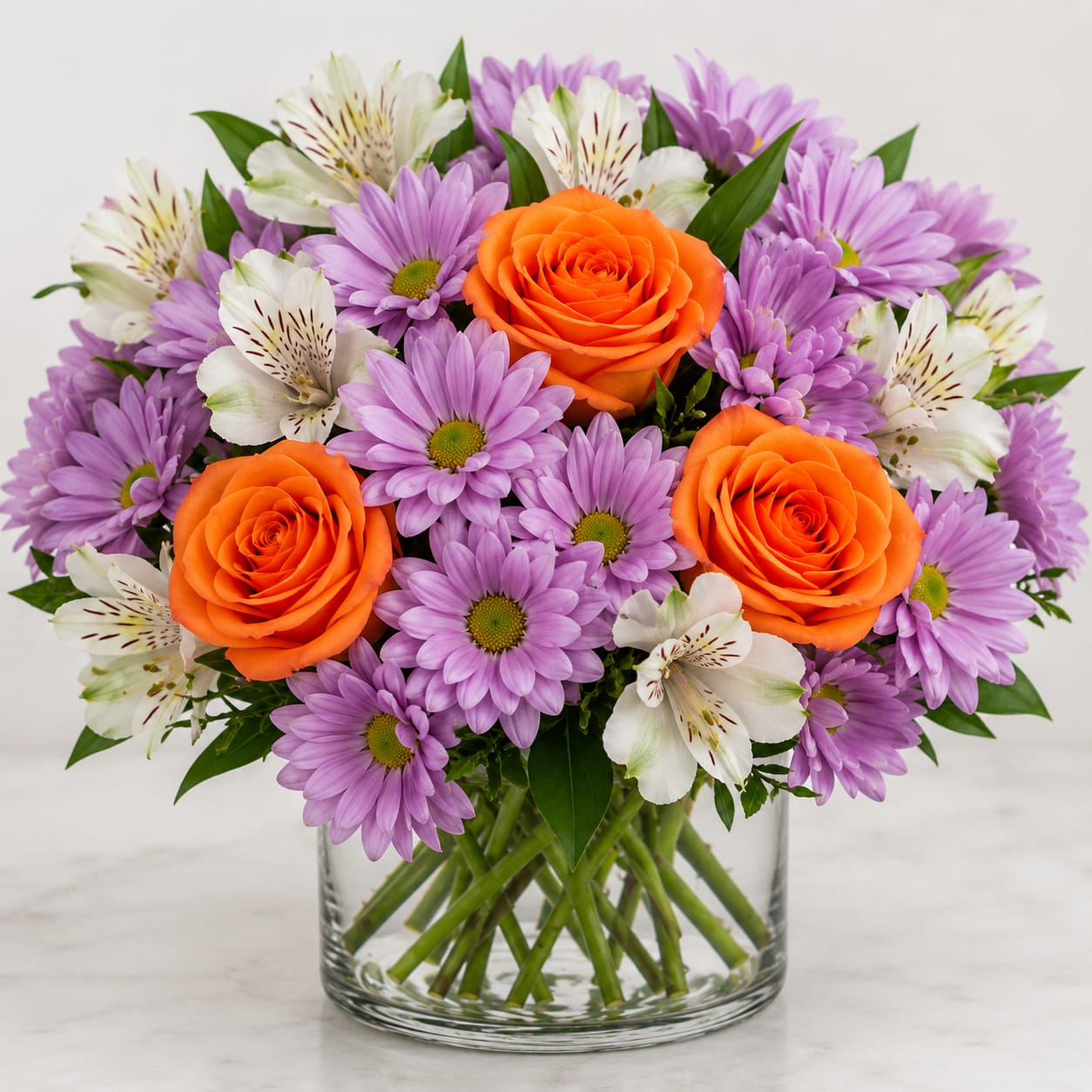 Citrus Lavender Delight - Bright, cheerful, and perfectly balanced — Citrus Lavender Delight blends soft elegance with vibrant energy. This compact, full arrangement features a lush base of lavender daisy mums, creating a soft garden texture, accented by three radiant orange roses that bring warmth and bold contrast. White alstroemeria adds a fresh, airy touch, enhancing fullness and giving the design a clean, polished finish.  Designed in a clear glass cylinder, this arrangement delivers a modern, high-impact look that photographs beautifully and sells easily across all platforms. The color combination makes it especially eye-catching for everyday occasions while still feeling premium.