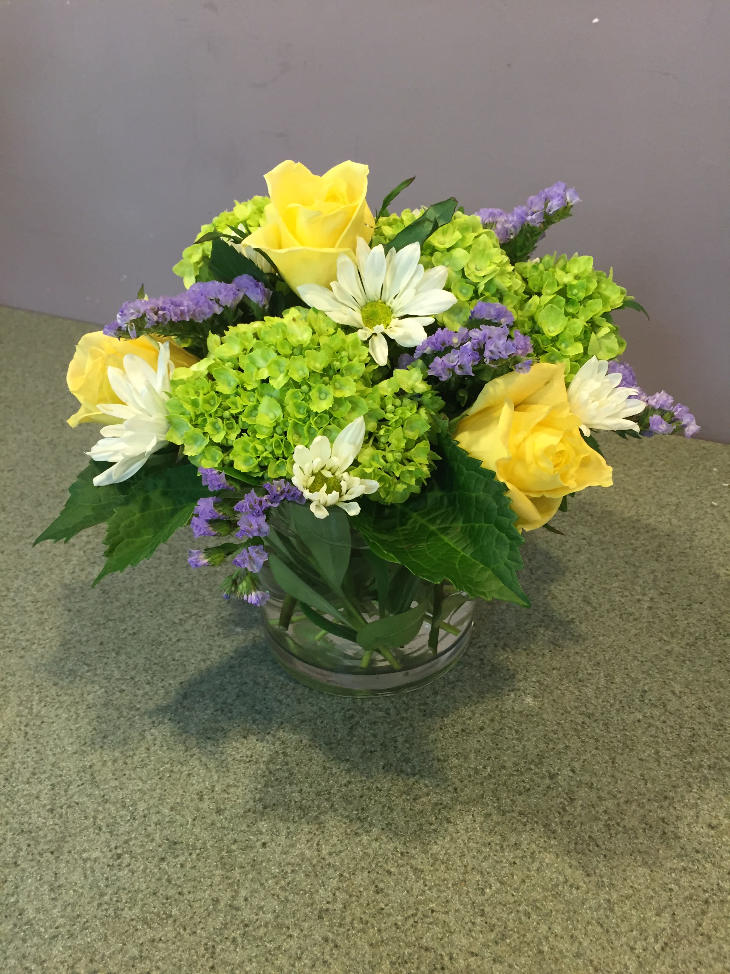 Yellow roses and white daisies in a glass bowl with green hydrangeas and purple filler flowers