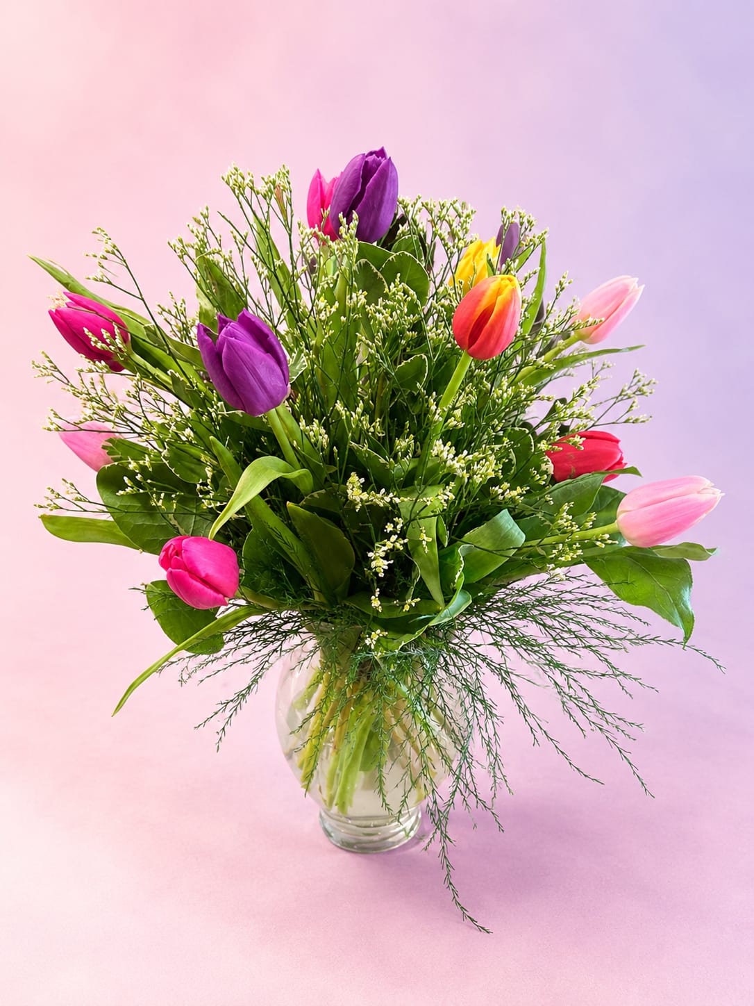 Tulip Garden Whimsy - Fresh, airy, and full of springtime charm, Tulip Garden Whimsy features a vibrant mix of colorful tulips nestled among lush greenery and delicate textural accents. Shades of pink, purple, coral, and sunshine yellow create a playful yet elegant palette, while wispy filler and flowing greenery give the arrangement a soft garden-inspired feel. Designed in a classic clear glass vase, this bouquet is perfect for celebrating spring, birthdays, Mother’s Day, or simply brightening someone’s day with cheerful seasonal beauty