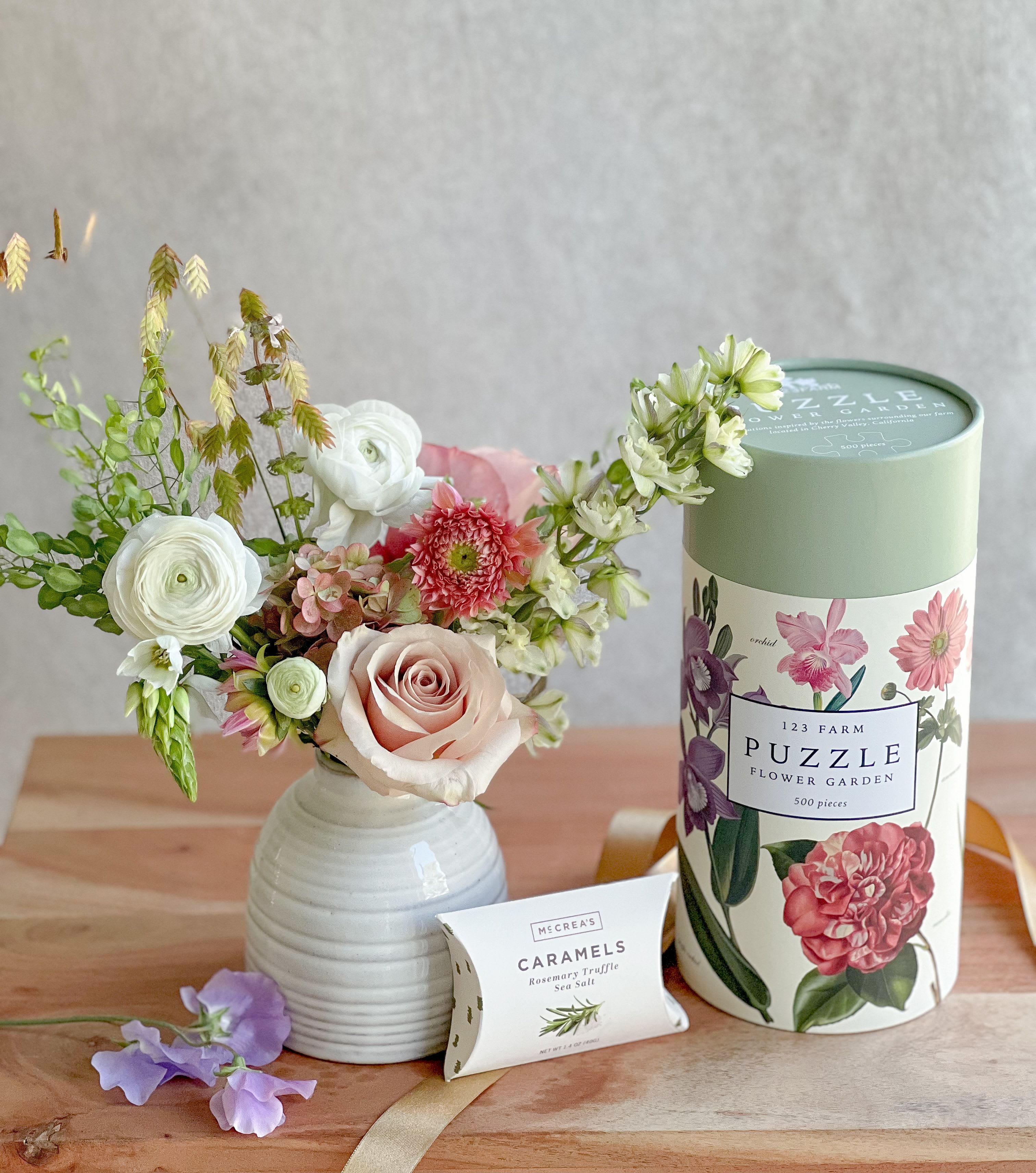 Mixed flower arrangement in a white vase beside a floral puzzle tin and caramel box