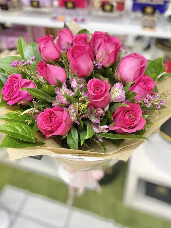 Hand-tied bouquet of bright pink roses with light pink accent flowers wrapped in patterned kraft paper.