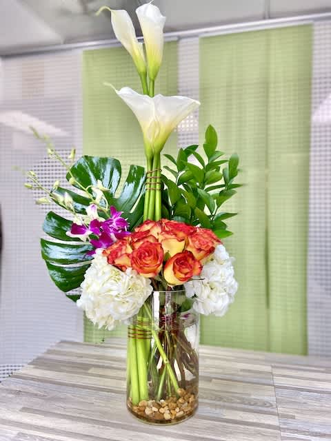 Tall arrangement of white calla lilies, orange roses, hydrangeas, and orchids in a glass vase with stones.