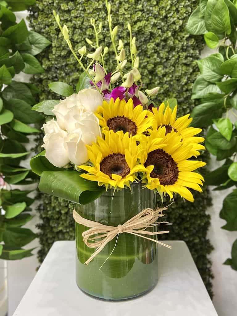Arrangement of sunflowers, white roses and orchids in a glass vase wrapped with a green leaf and raffia bow