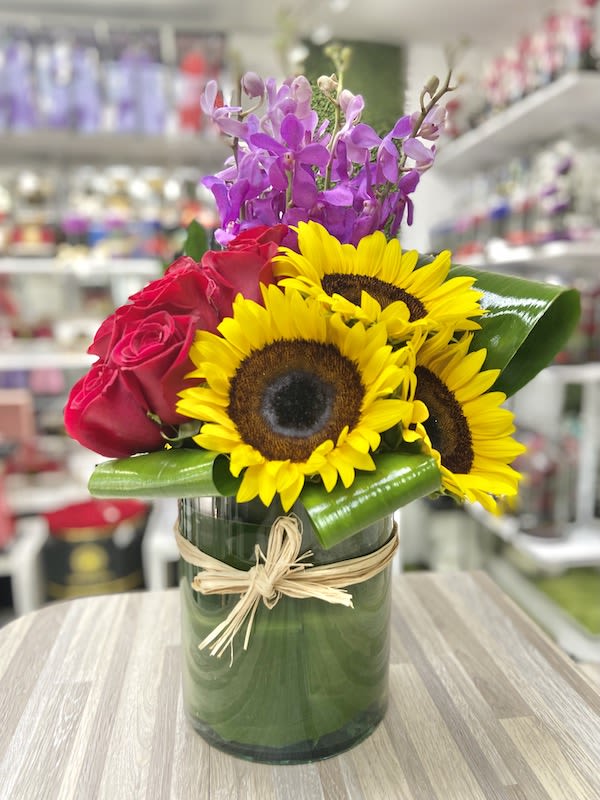Mixed arrangement of sunflowers, red roses, and purple orchids in a glass cylinder vase with raffia bow