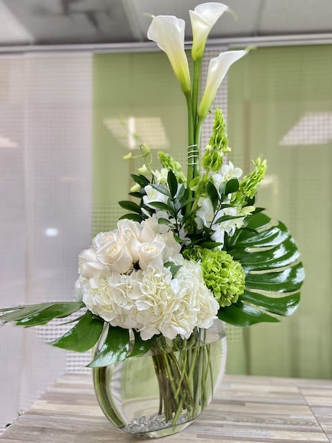 Tall white calla lilies with white roses and hydrangeas in a clear glass vase.