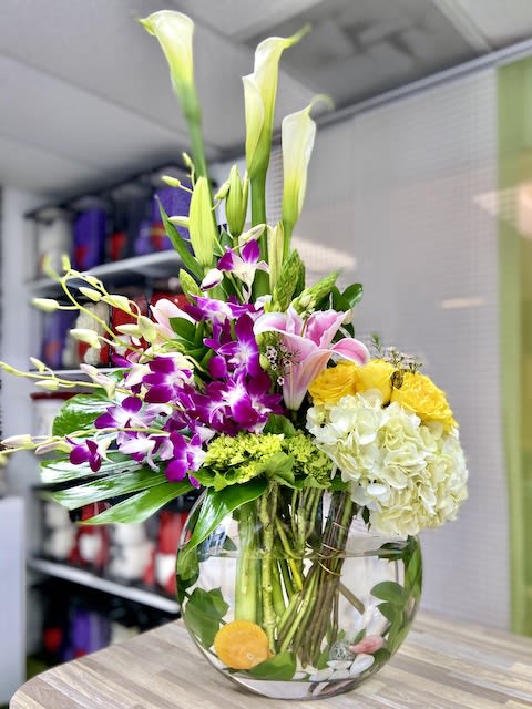 Tall mixed arrangement with white calla lilies, purple orchids, pink lilies, and yellow blooms in a round glass vase