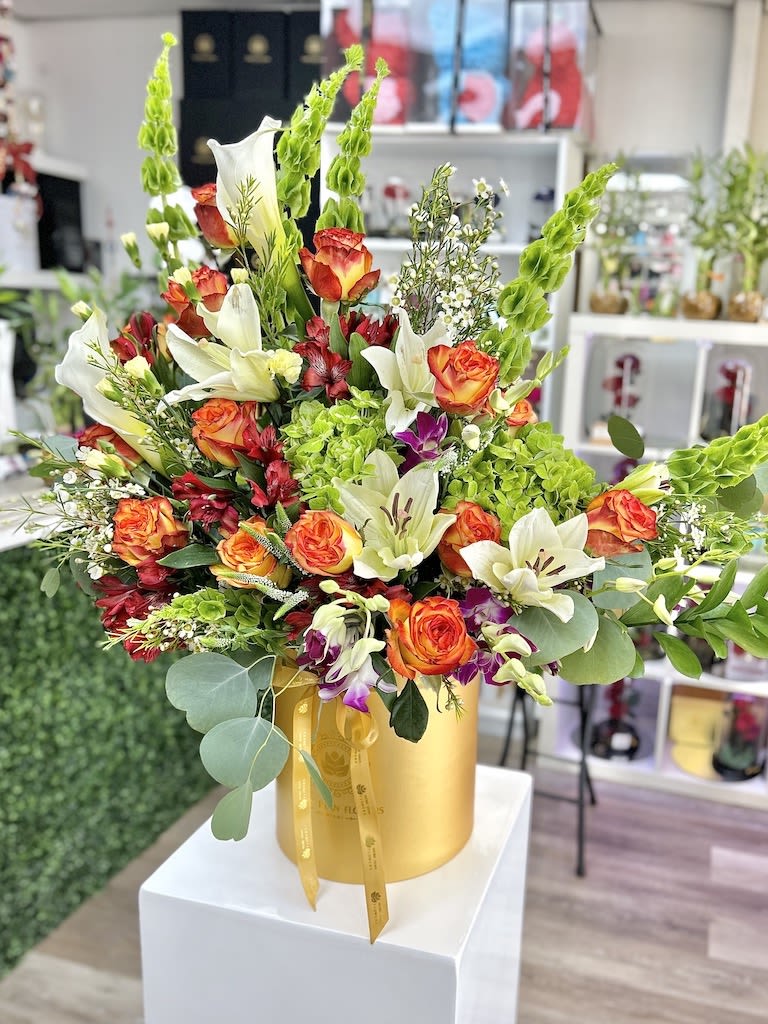 Tall arrangement of orange roses and white lilies in a gold hatbox-style container.