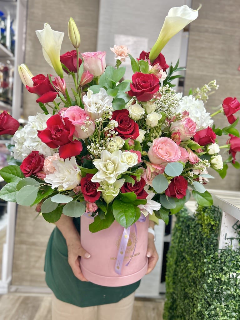 Tall arrangement of red and pink roses with white blooms and calla lilies in a pink hatbox container