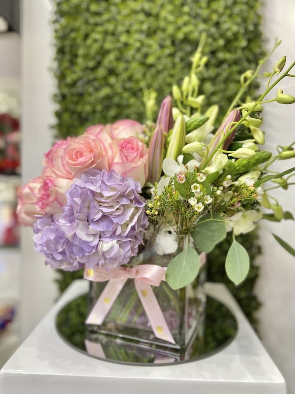Pastel arrangement of pink roses, lavender hydrangea, and lilies in a glass cube vase with a pink ribbon.