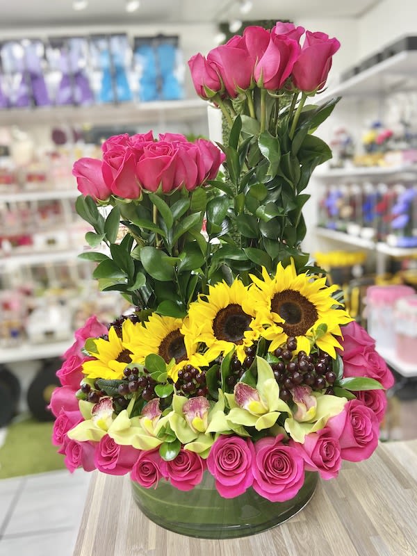 Structured arrangement of hot pink roses, yellow sunflowers, green orchids and grapes in a low glass vase