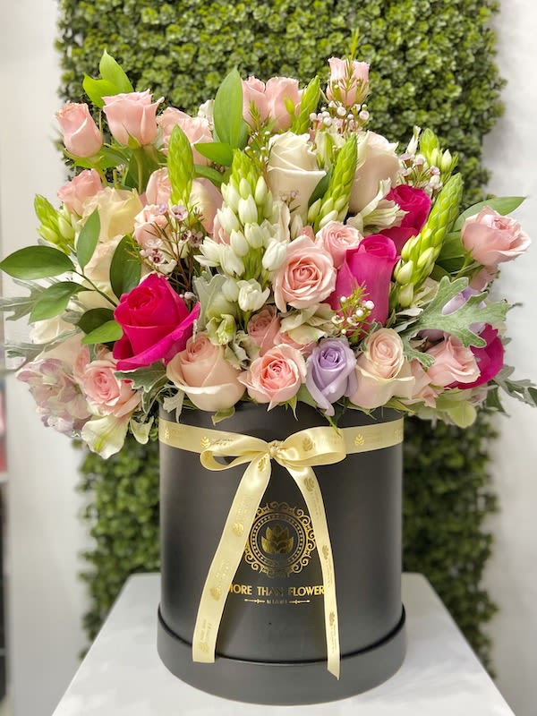 Round hatbox of mixed pink, cream, and purple roses with white accent flowers and a yellow ribbon bow.
