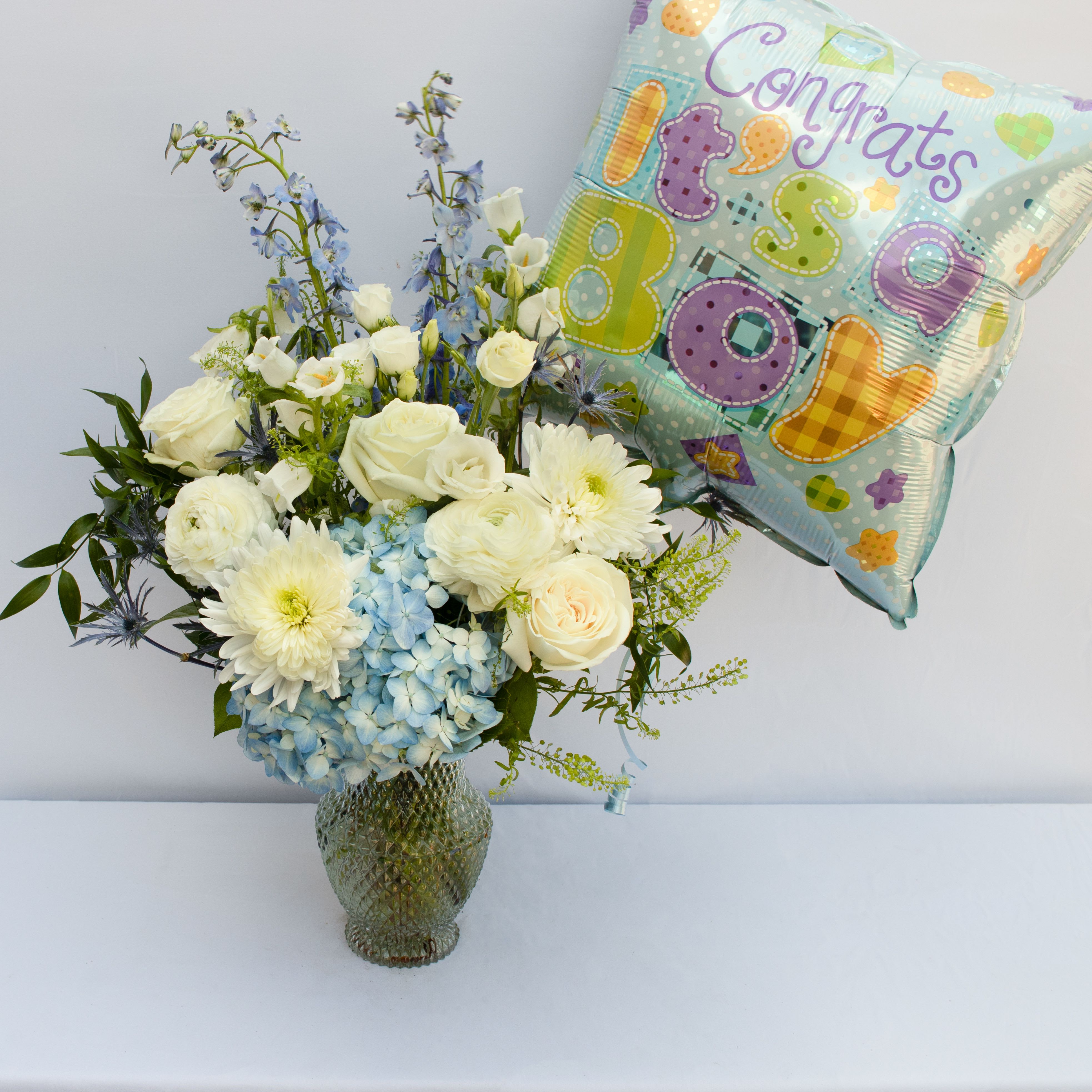 IT'S A BOY!  - BLUE SKY ARRANGEMENT WITH AN "IT'S A BOY BALLOON ADDED" A SOFT MIXTURE OF WHITE AND BLUES TO INCLUDE SOME OF THESE FLOWERS SNAPDRAGONS, HYDRANGEA, ROSES, GERBERAS, LISIANTHUS, RANUNCULUS, CAMPANULA, ALSTROMERIA, MUMS THISTLE ACCENTED WITH GREEN DRAGON ARRANGEMENT MADE IS ABOUT 14HX8W.  ARRANGEMENT IS IN A SOFT LIGHT BLUE BUBBLE TEXTURED VASE.  