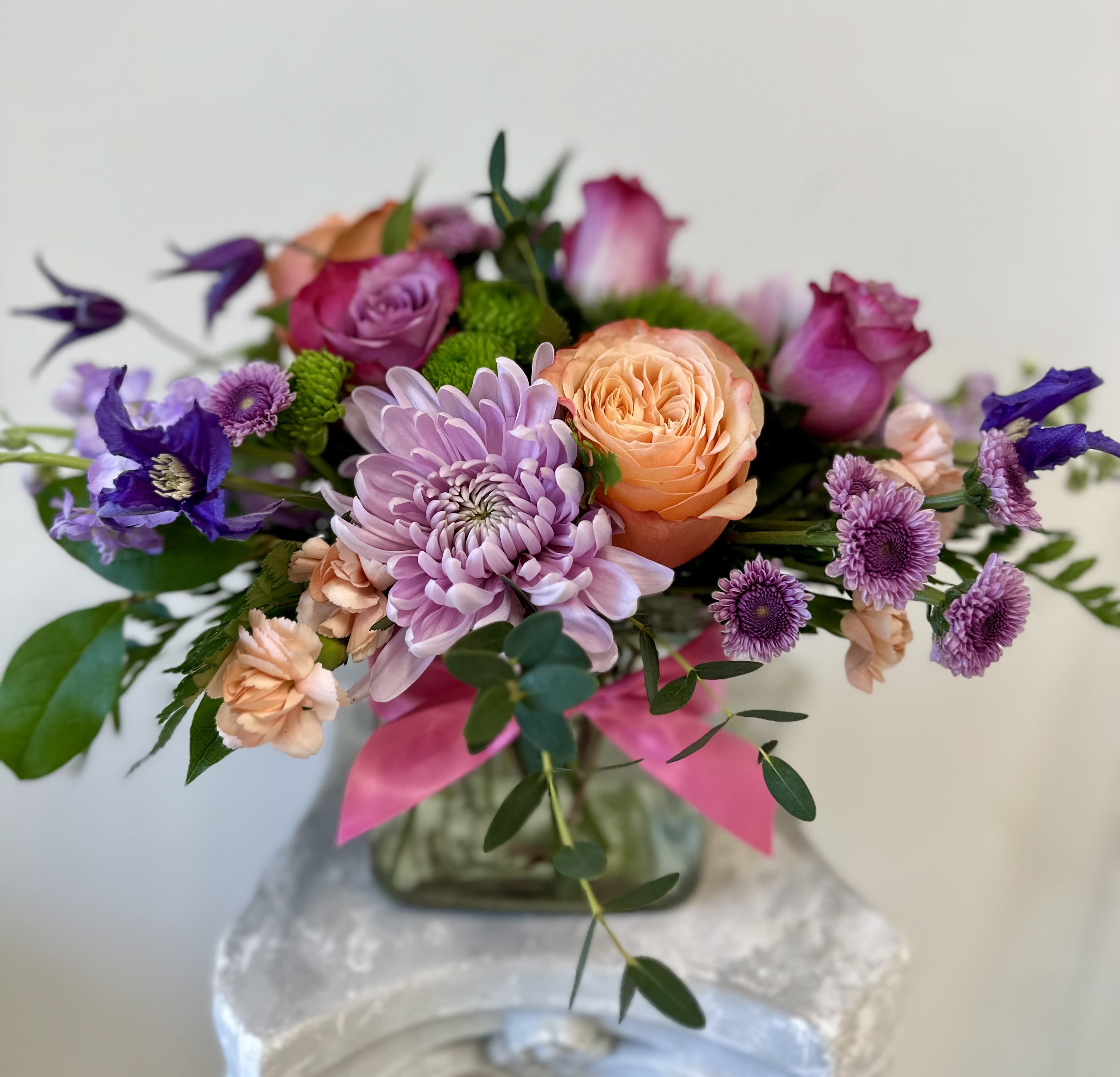 Low arrangement of purple mums, peach roses, and mixed blooms in a glass vase with a pink ribbon.