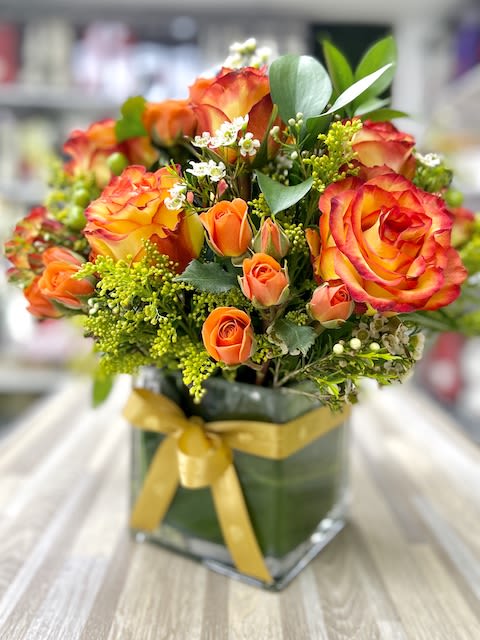 Orange and yellow roses with small white flowers in a leaf-lined square glass vase with a gold ribbon bow.