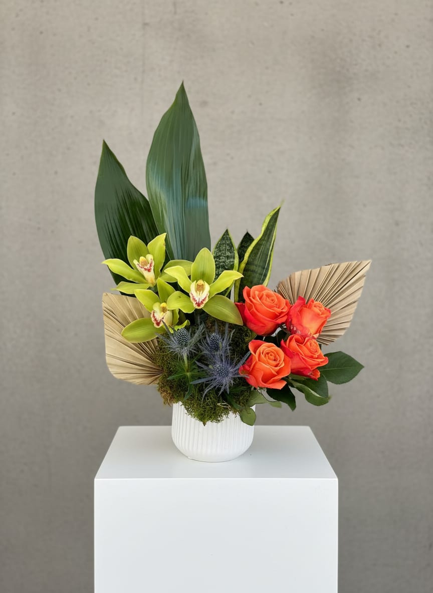 Modern arrangement with green orchids, orange roses, palm fans and large green leaves in a white vase.