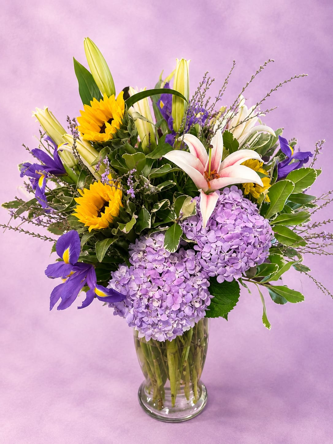 Golden Lavender - Bright yet elegant, Golden Lavender combines cheerful sunflowers with soft lavender hydrangea, graceful lilies, and vibrant blue iris blooms for a stunning blend of color and texture. Accented with lush greenery and delicate wispy filler, this arrangement brings together warm golden tones and cool lavender hues in a fresh, garden-inspired style. Designed in a classic clear glass vase, it’s a beautiful choice for birthdays, celebrations, encouragement, or simply brightening someone’s day with something unique and uplifting.